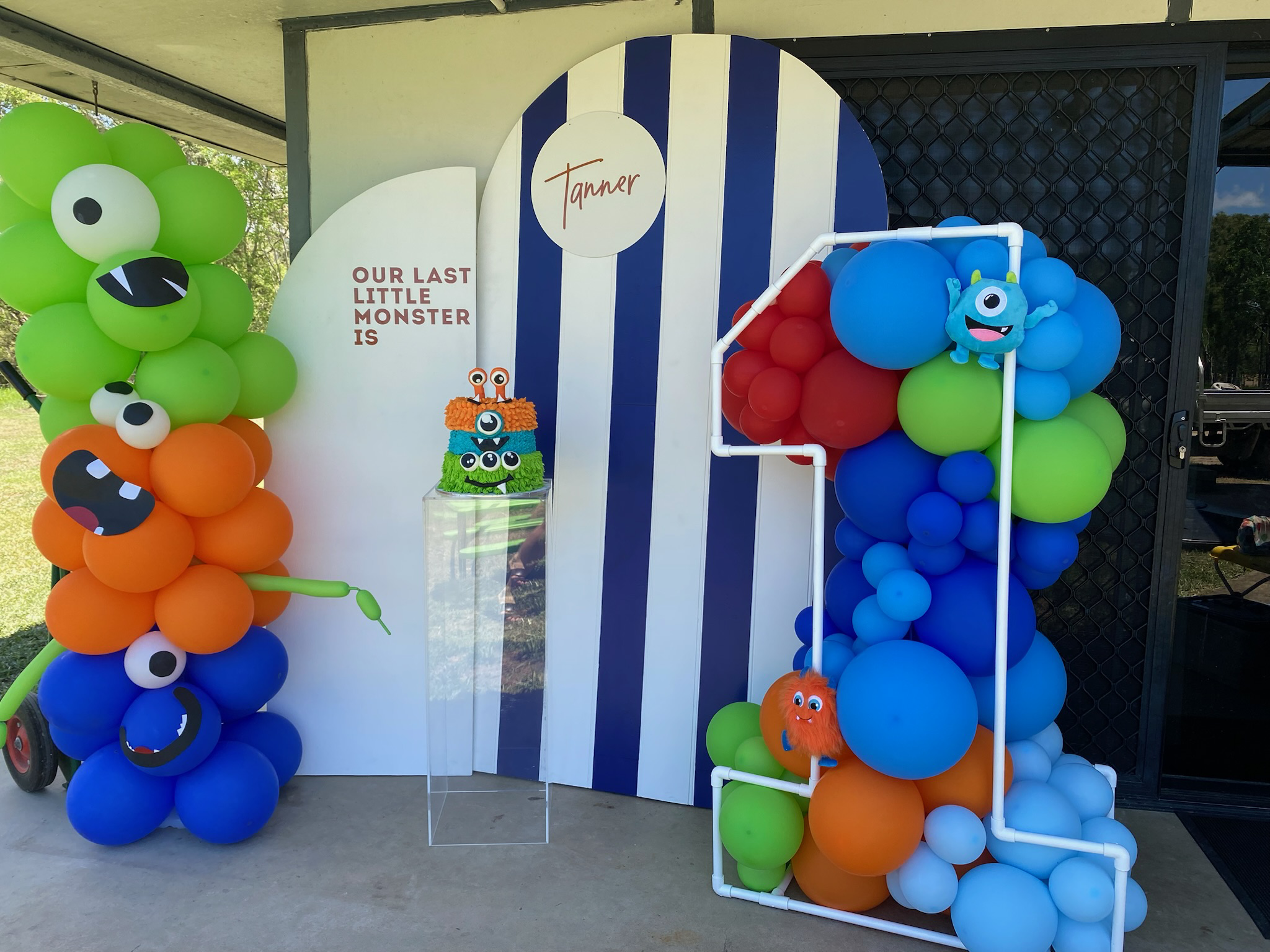 Colorful balloon display with cartoon monster decorations for a birthday party, with a striped backdrop and a sign that reads "Our last little monster is Tanner."
