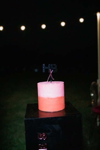 Pink birthday cake with an 'HB' topper on a black table outside at night, with string lights overhead.