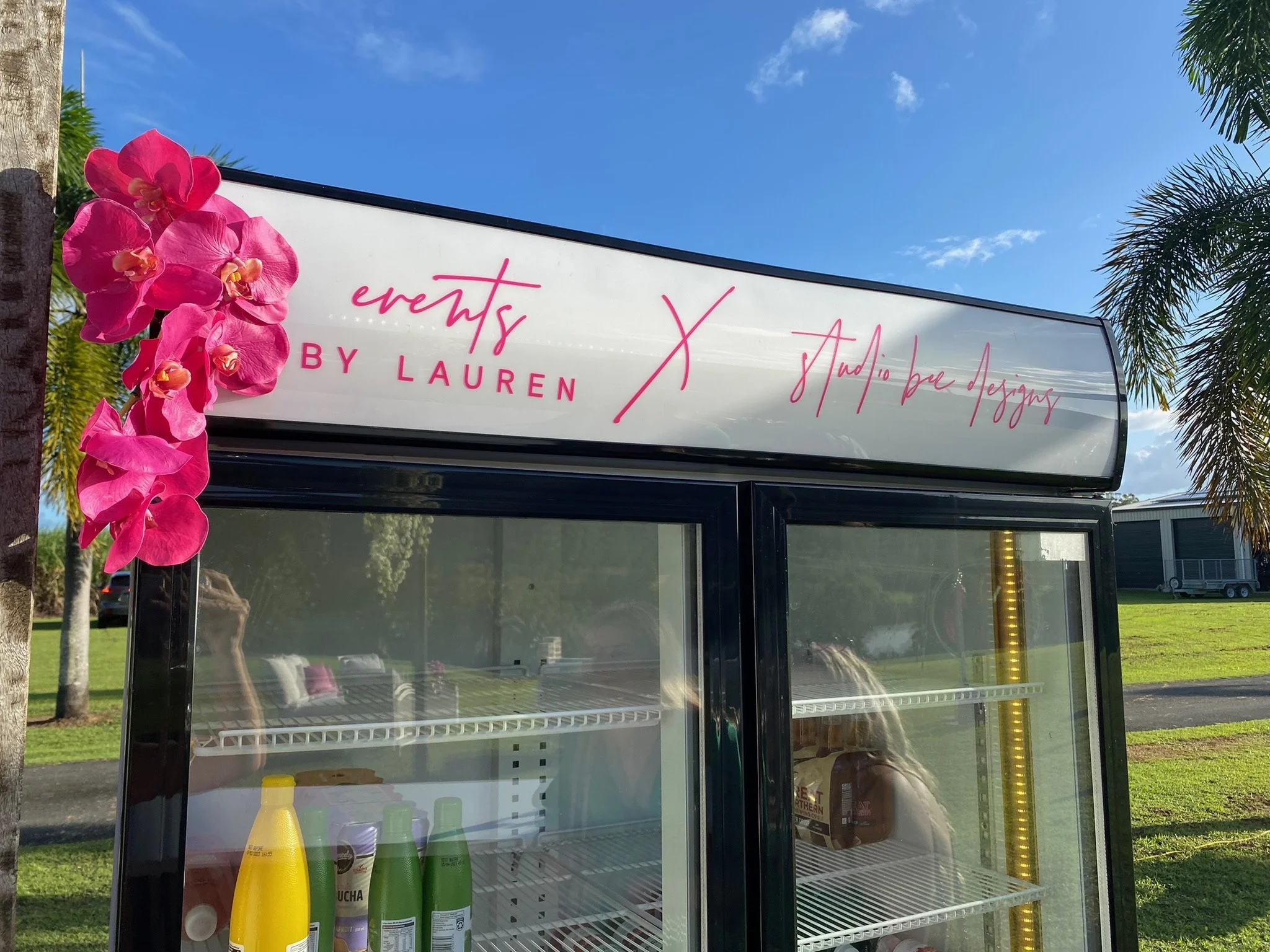 A black and glass display refrigerator with pink orchids attached on the top left corner. The signage on top reads "Events by Lauren x Studio Bae Designs" in pink script. The refrigerator contains some bottles of beverage and is set outdoors on grass