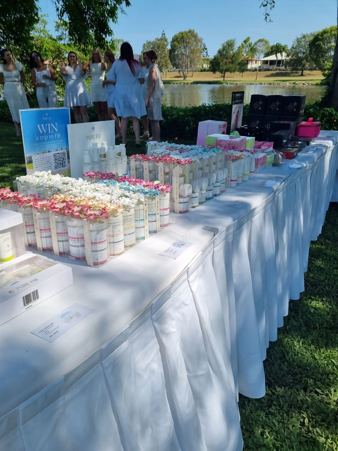 Table displaying skin care products at an outdoor event near a lake with a group of women in white dresses in the background.