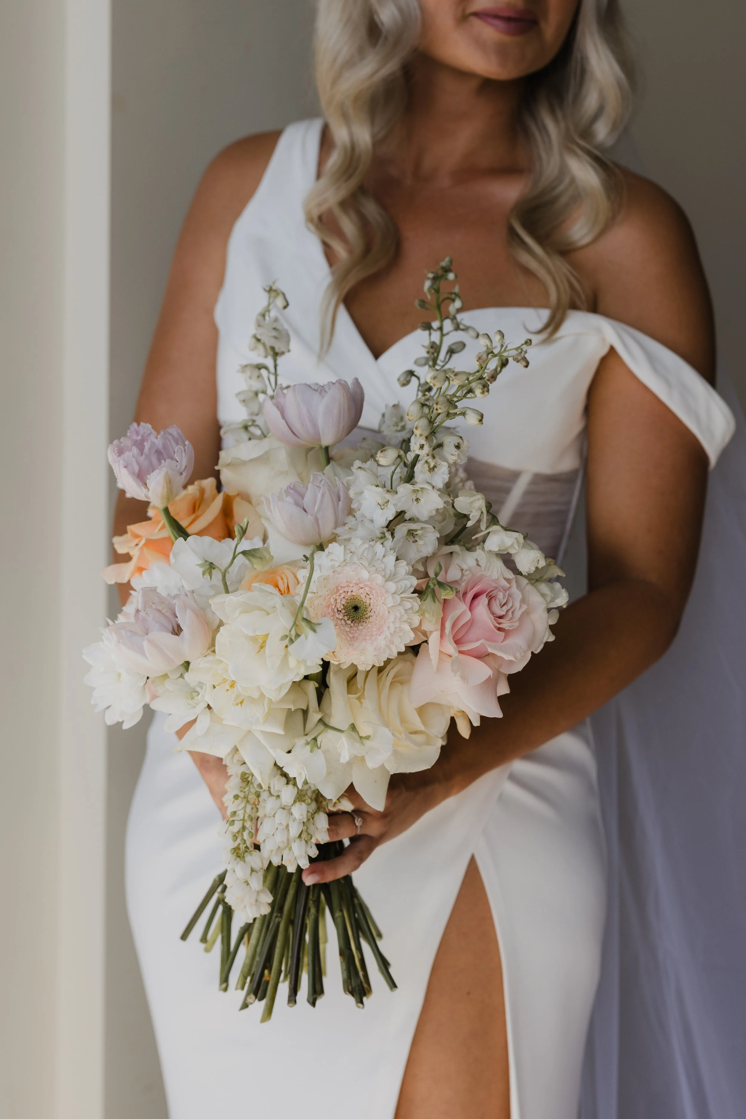 A woman in a white dress holding a bouquet of pastel-colored flowers, including roses, tulips, and other blooms, with a slit in her dress revealing her leg.