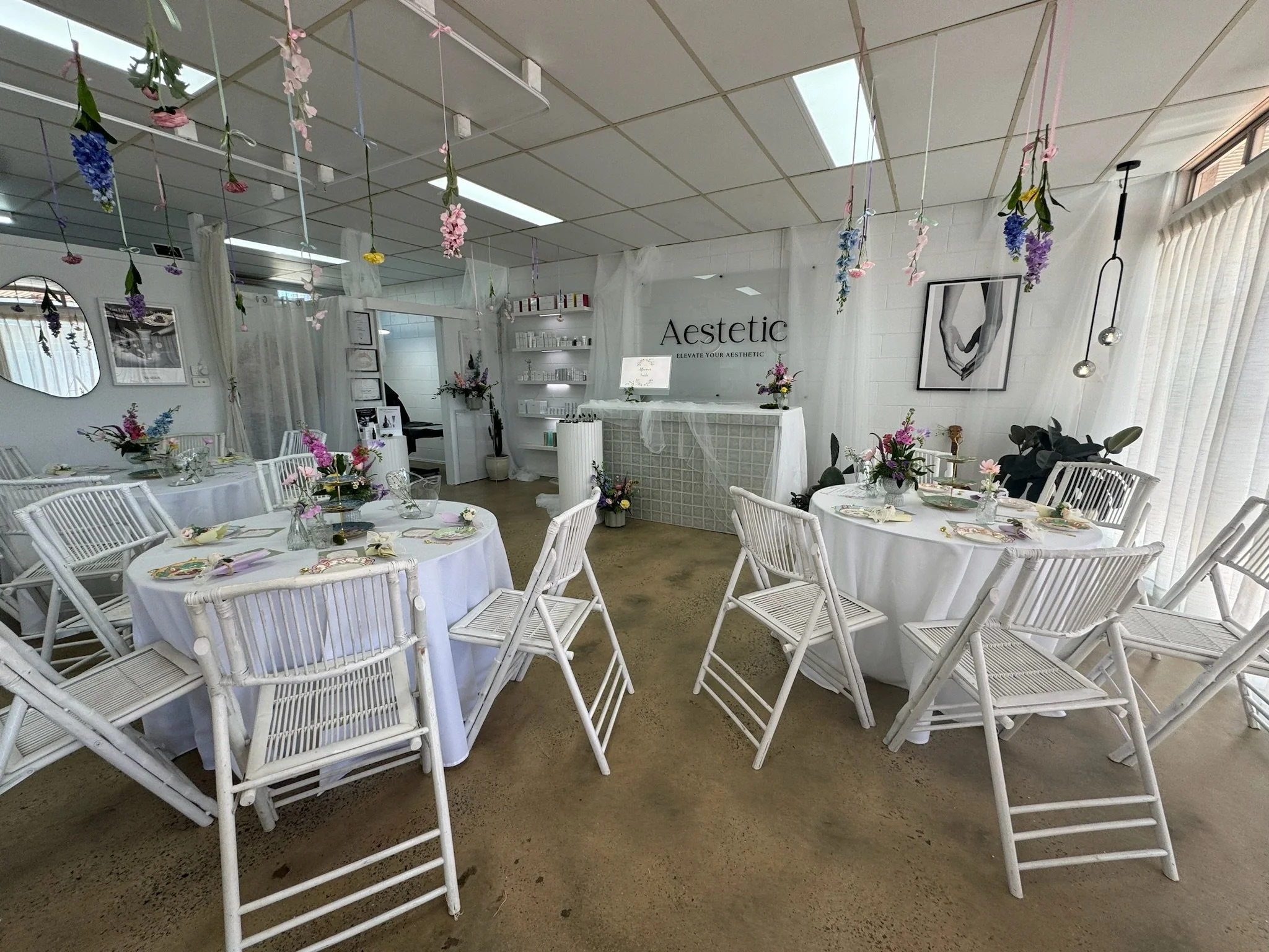 Elegant event space with white round tables dressed in white tablecloths, decorated with floral centerpieces, set with plates, cups, and napkins, surrounded by white chairs. Hanging flower arrangements from the ceiling and black and white wall art en