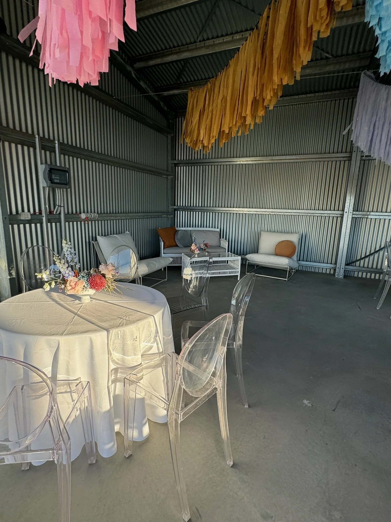 Indoor event space with metal walls and ceiling, decorated with hanging pastel-colored paper tassels, featuring a round table with a floral centerpiece, clear acrylic chairs, and a seating area with white and gray couches and pillows.