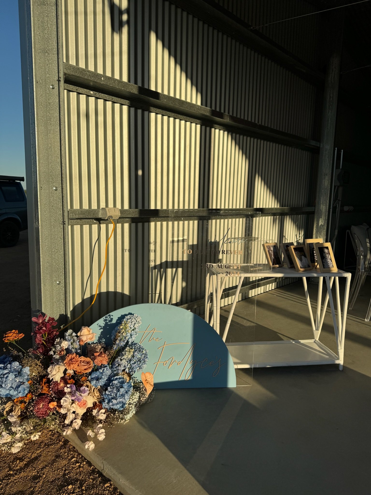 Flowers, a sign for 'The Fordace's', and framed photos on a white table against a metal wall with sunlight and shadow patterns.