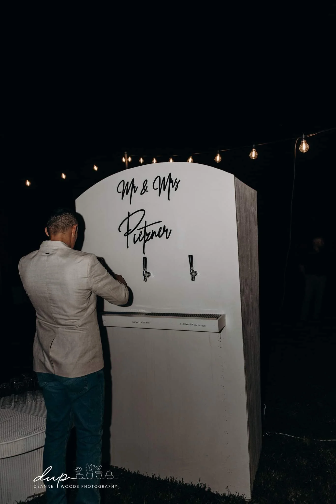 A man in a beige jacket filling a drink from a tap at a wedding reception with a sign that says "Mr & Mrs Pitetner" and string lights overhead.