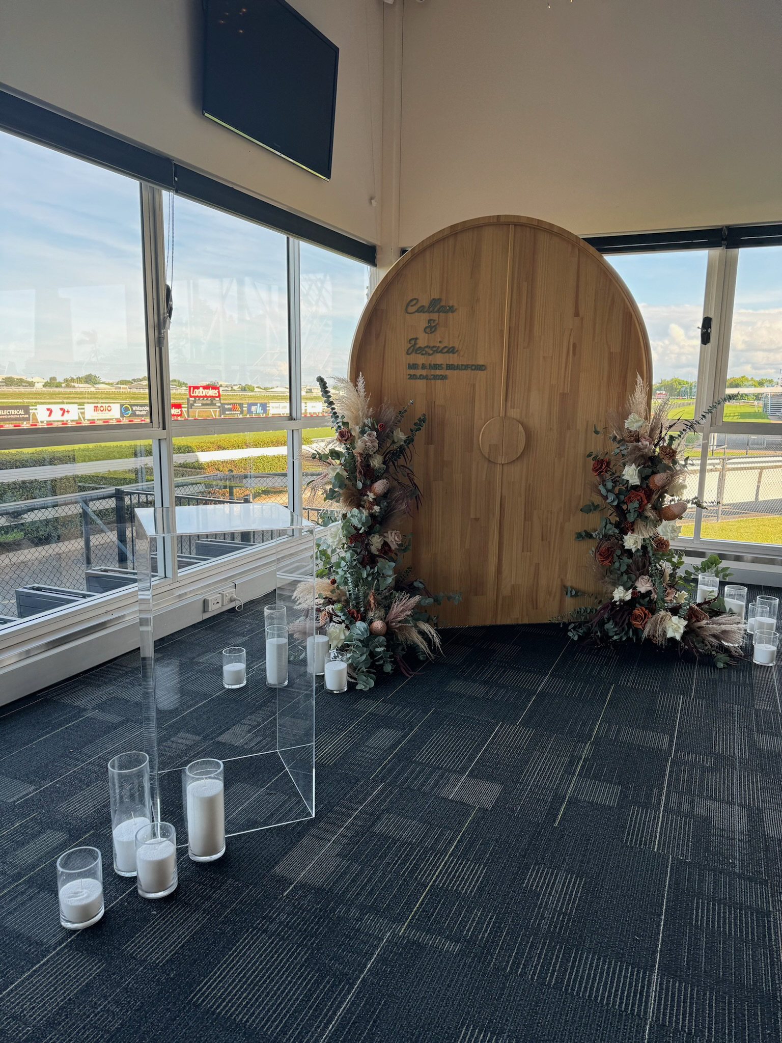 Wedding backdrop with wooden arch, floral arrangements, candles, and a clear acrylic lectern on a dark carpeted floor at a racetrack with large windows showing the outdoors.