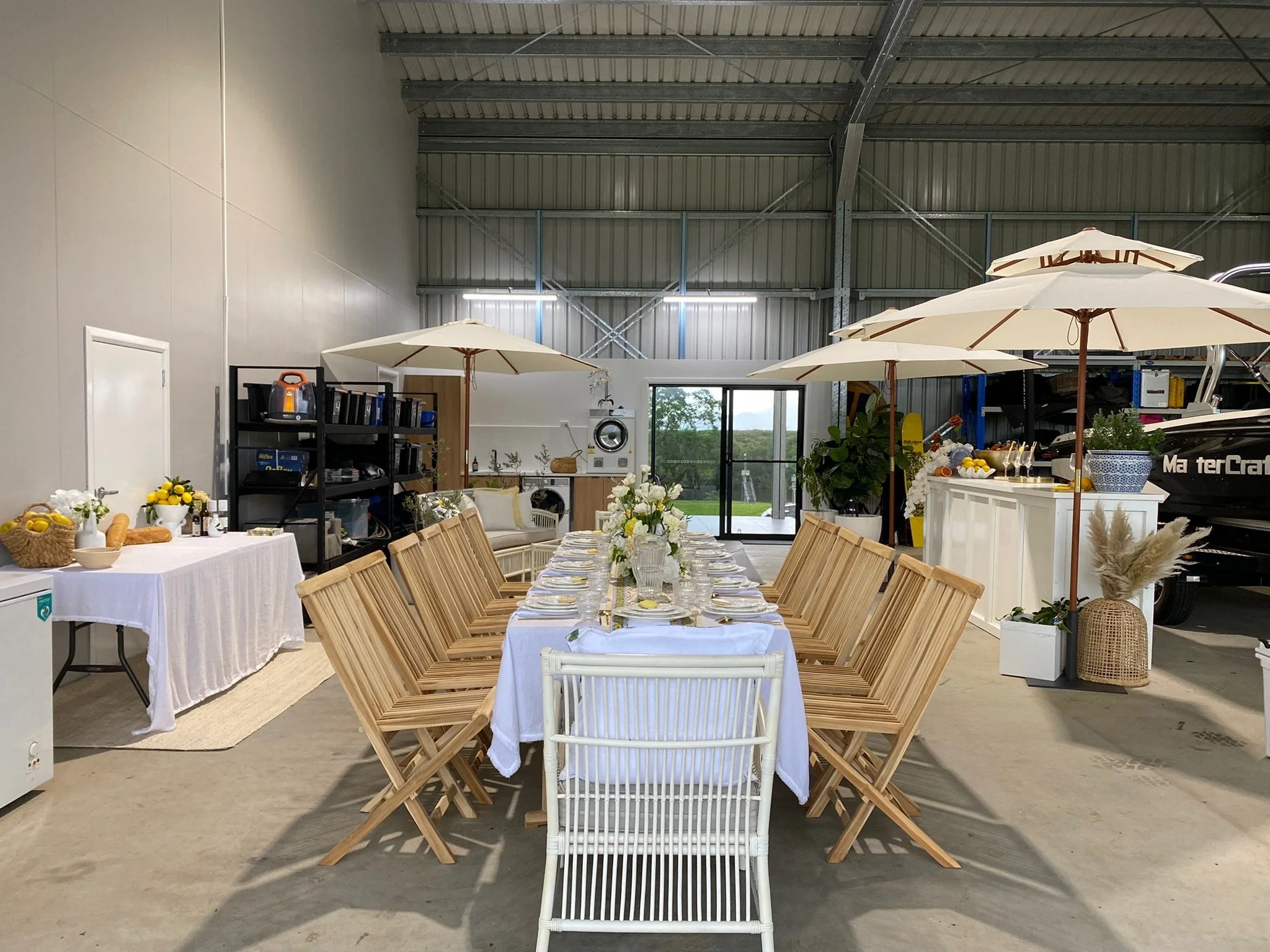 Indoor party setup featuring a long dining table with white tablecloth, surrounded by wooden chairs, decorated with floral arrangements, umbrellas, and tableware. There's a lounge area in the back, a side table with food and drinks, and a large boat 