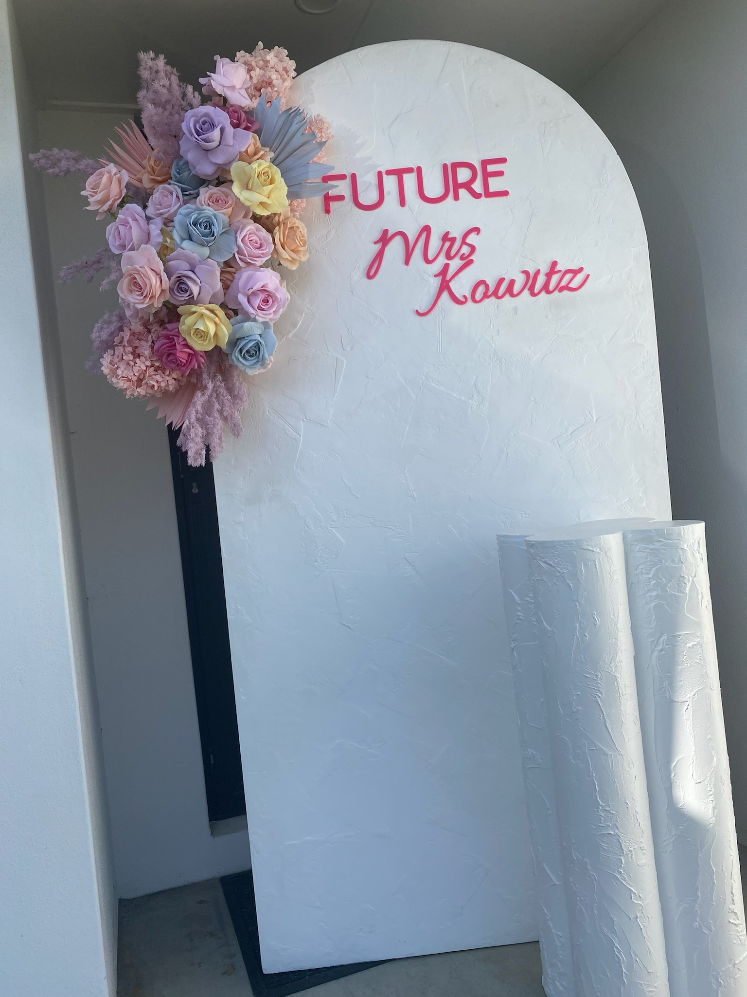 A white, curved backdrop with pink text that reads 'FUTURE Mrs Kowitz'. A floral arrangement featuring pastel pink, purple, yellow, and blue roses, with additional pink and purple pampas grass, decorates the top left corner of the backdrop.