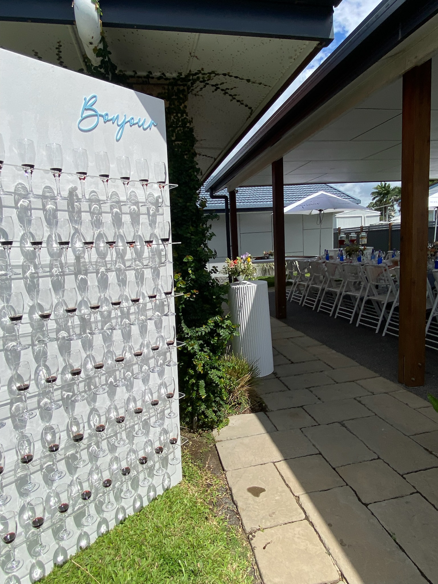 Outdoor celebration setup with a white board displaying many small wine glasses, some filled with red wine, under a shaded patio with tables and chairs.