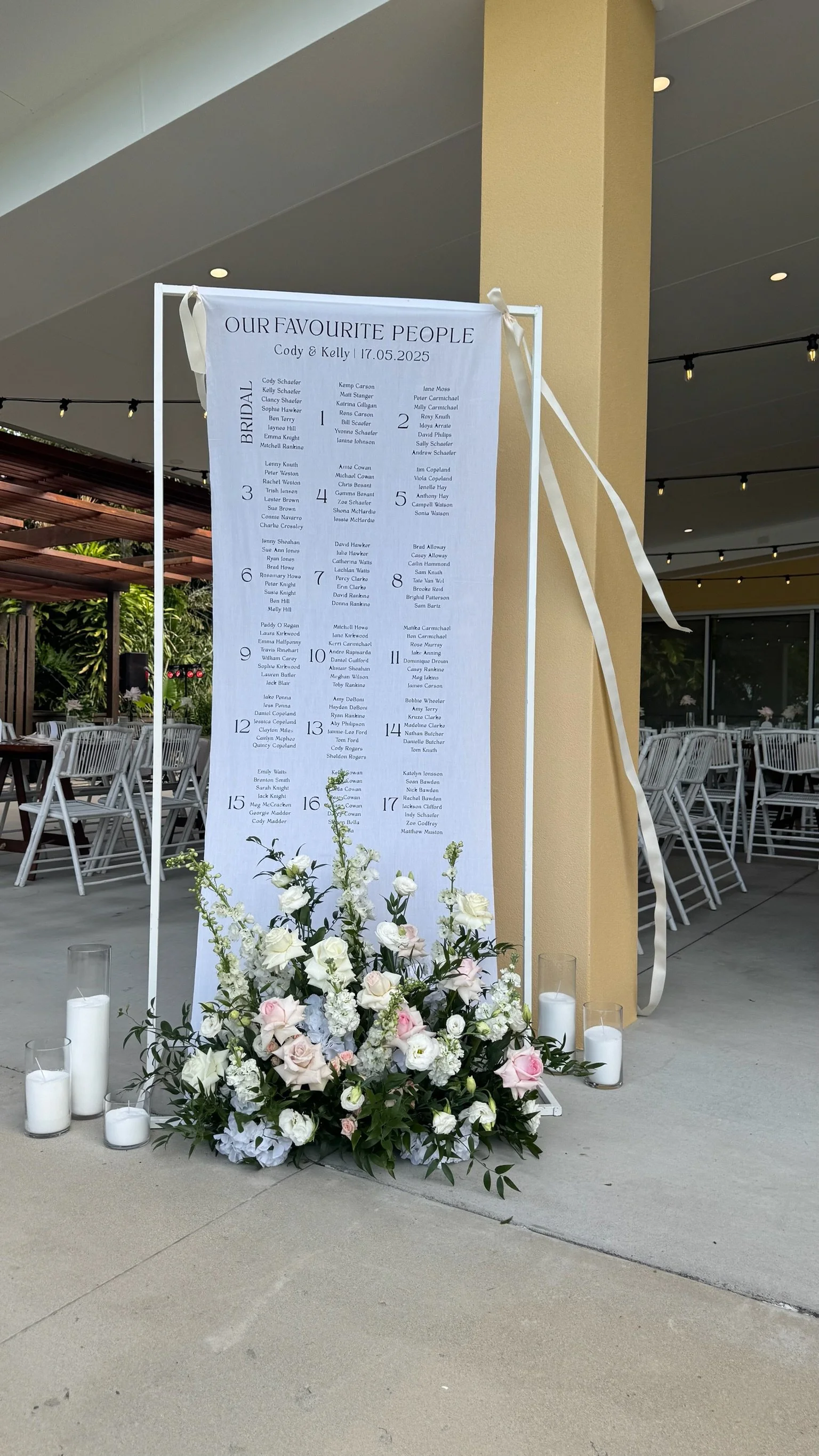 Wedding seating chart sign decorated with white and pink roses and candles, with tables numbered and names listed for the wedding of Cody and Kelly on May 17, 2025.