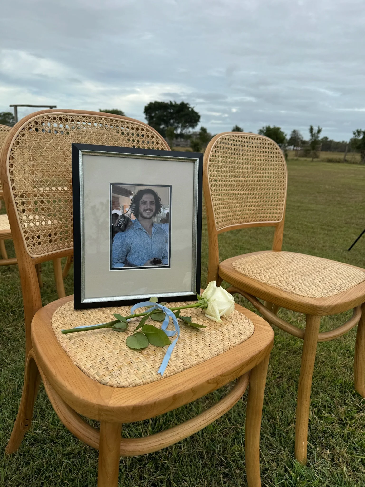 A framed black-and-white photograph of a man with long curly hair smiling, holding a beer bottle in his right hand, is placed on a wooden chair outdoors. On the seat of the chair, there is a white rose with a blue ribbon tied around its stem. The bac
