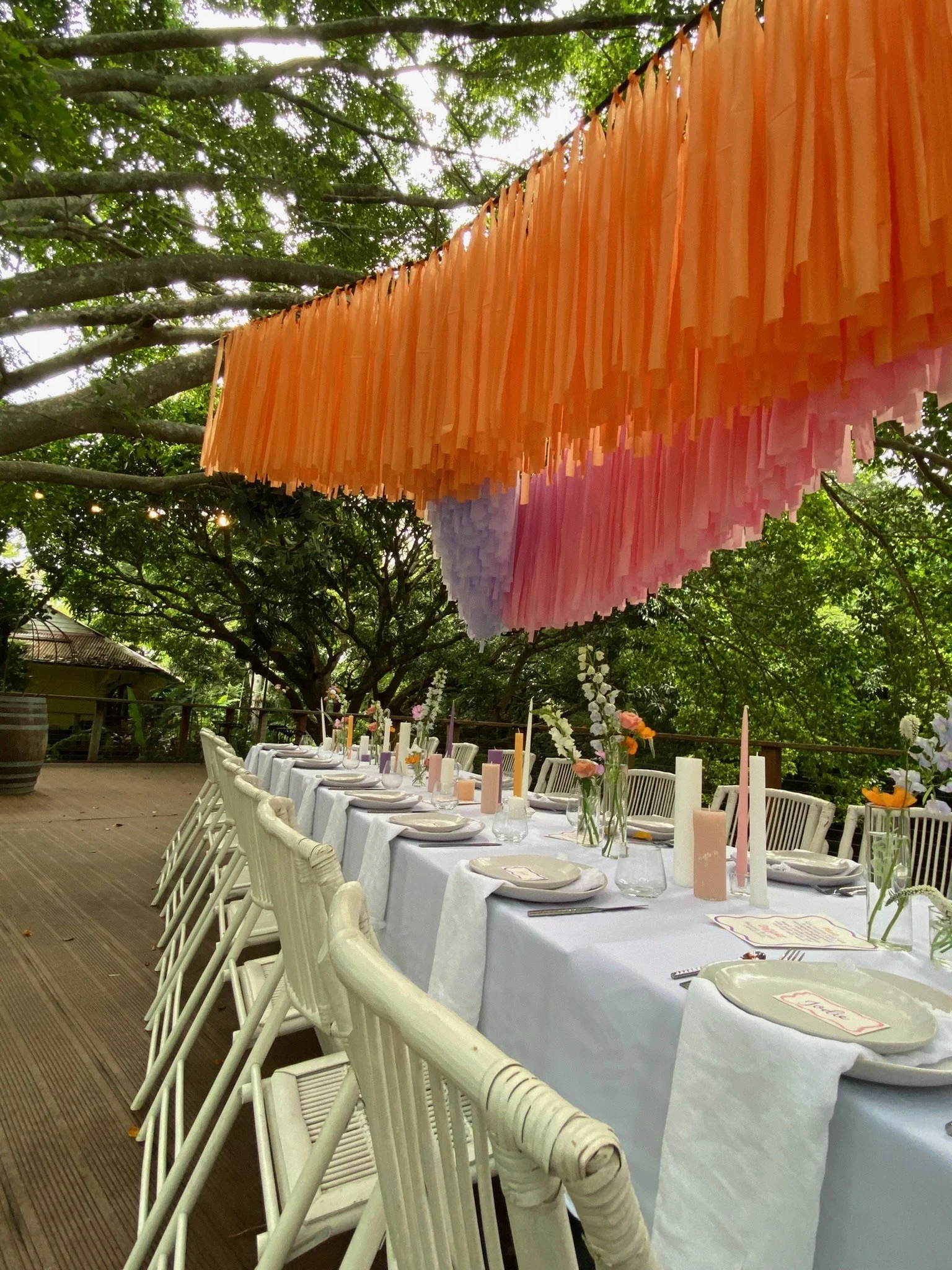 Elegant outdoor dining setup with a long white table, candles, and floral centerpieces, under a colorful paper fringe canopy, surrounded by green trees.