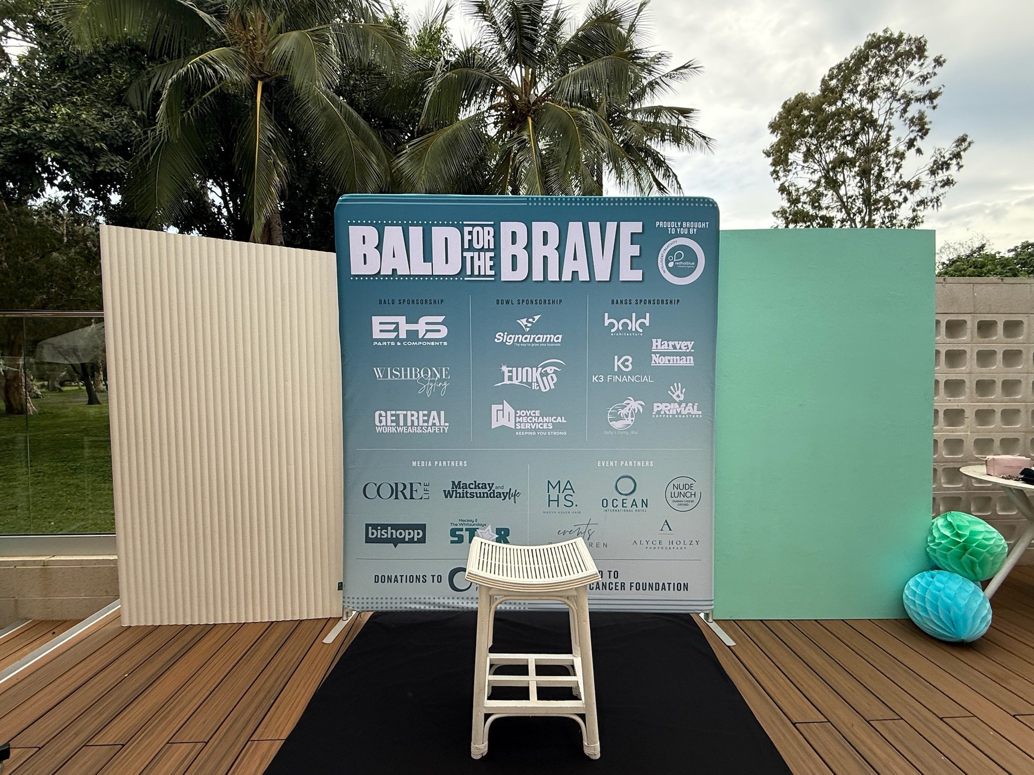 A display board with the title "Bald for the Brave" and sponsor logos at an outdoor event, with two decorative paper lanterns on the right side and a white stool in front of the board.