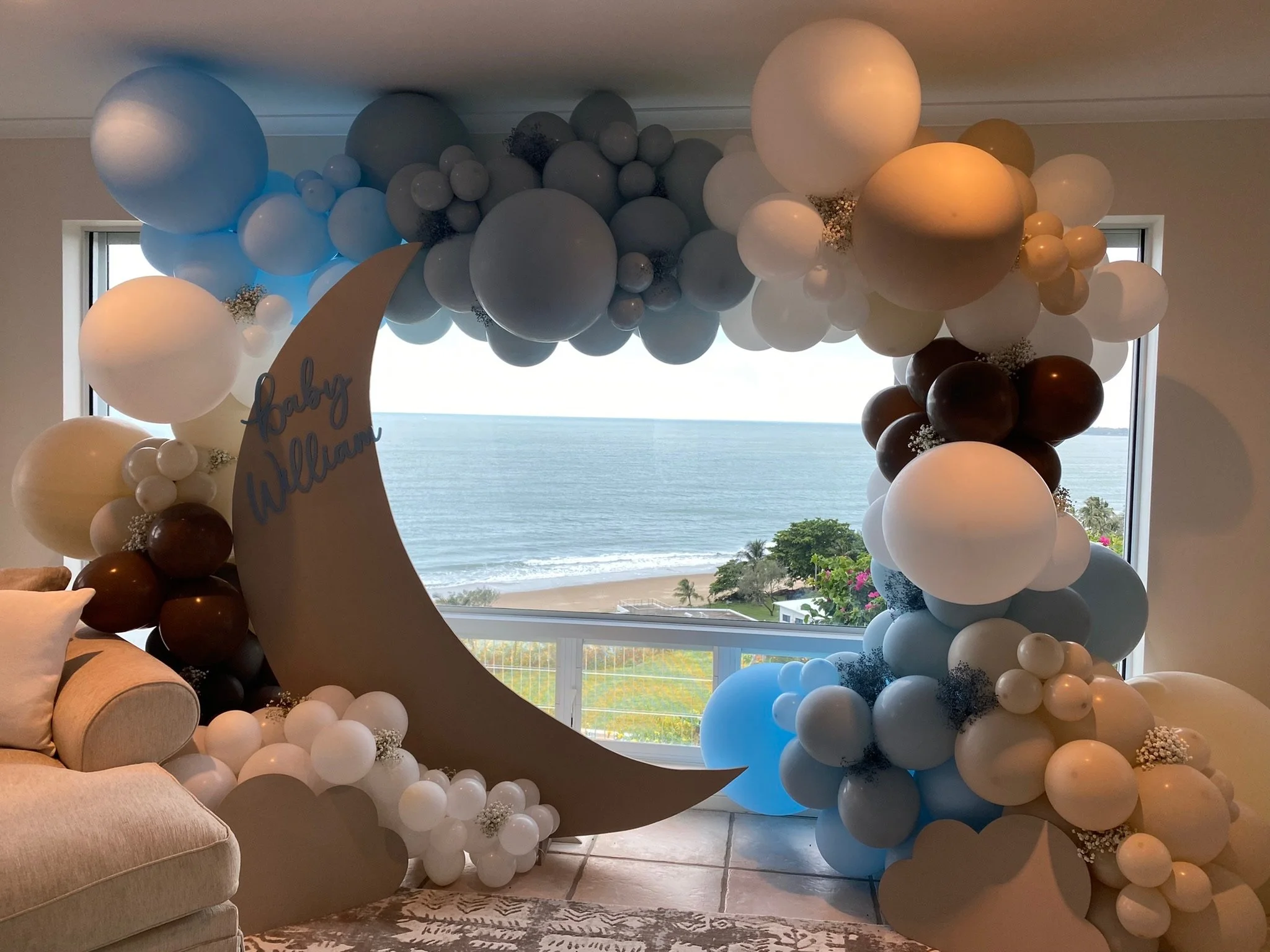 Decorative balloon arch with white, black, and beige balloons, crescent moon cutout with 'baby William' written on it, overlooking ocean view from inside a room.