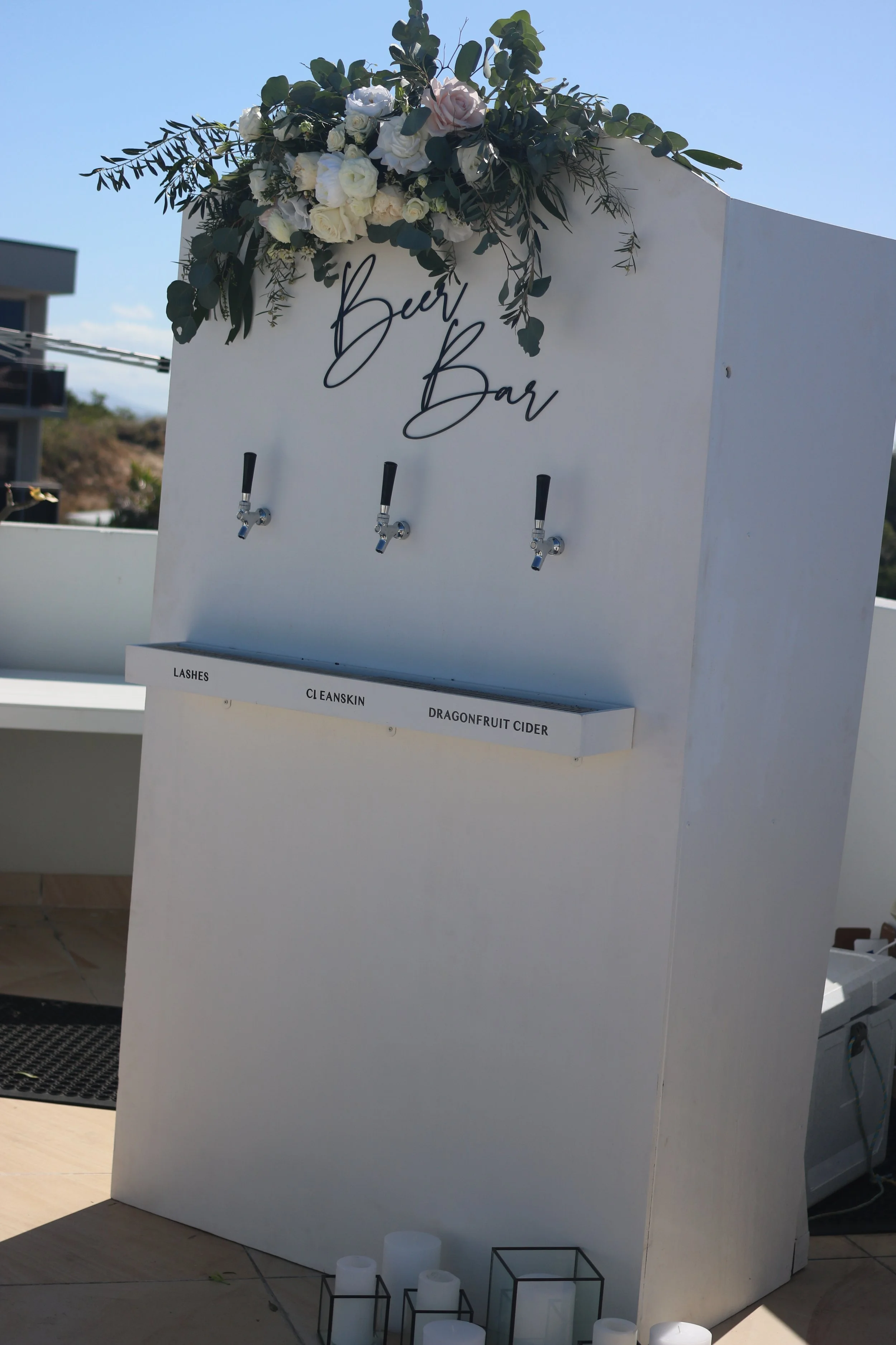 A beverage station with three taps labeled for beer, with a floral arrangement on top, and options listed below for lashes, cleanskin, and dragonfruit cider, set outdoors during the daytime.