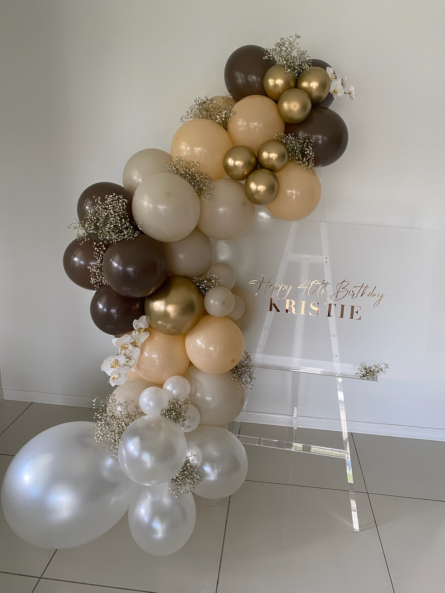 Balloon arrangement with white, cream, gold, and brown balloons, decorated with small white flowers, and a clear stand with a sign that reads "Happy 40th Birthday Kristie."