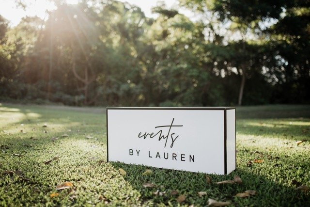 A sign on grass reading 'events by Lauren' in a park with sunlight filtering through trees.