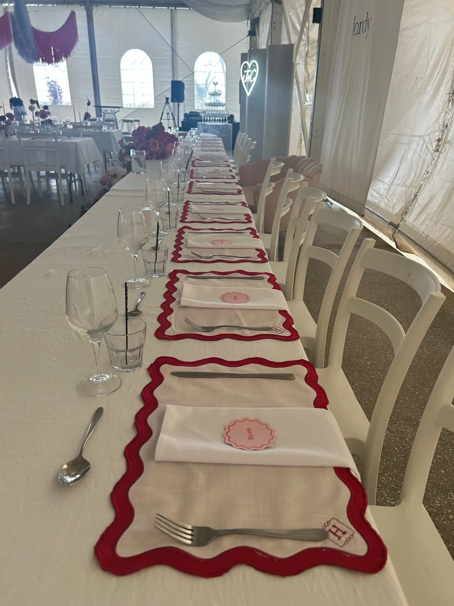 Wedding or event reception table set with white tablecloth, pink and red placemats, white napkins with pink labels, forks, knives, empty glasses, and small flower arrangements. Tent with windows and pink hanging decorations visible in the background.