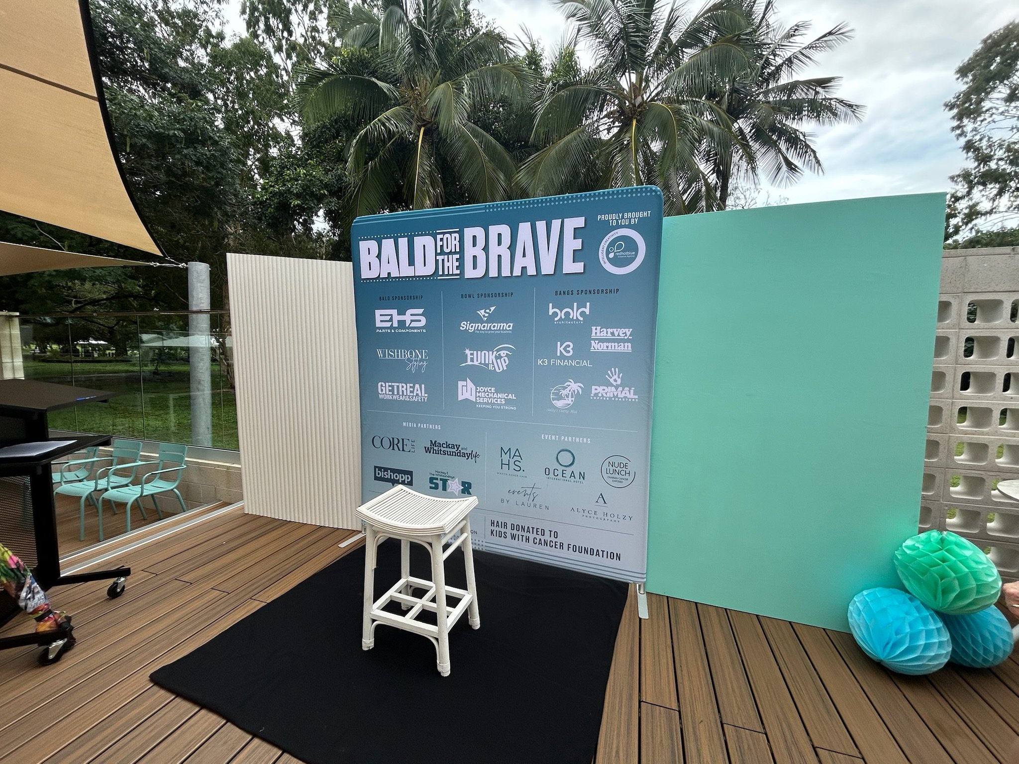 Event banner titled 'Bald for the Brave' on a sign with various sponsor logos, set up outdoors on a deck with palm trees in the background, alongside decorative paper lanterns in blue and green.