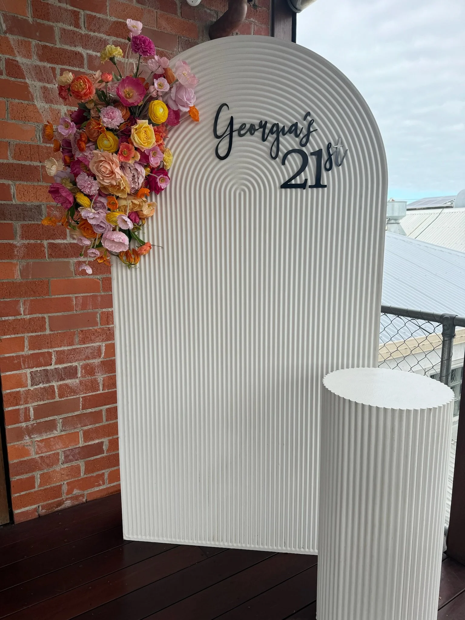 A decorative totem with a large white ribbed oval structure with black cursive and bold text that reads 'Georgia's 21st.' A floral arrangement of pink, yellow, orange, and purple paper flowers along the top left side of the white structure. A matchin