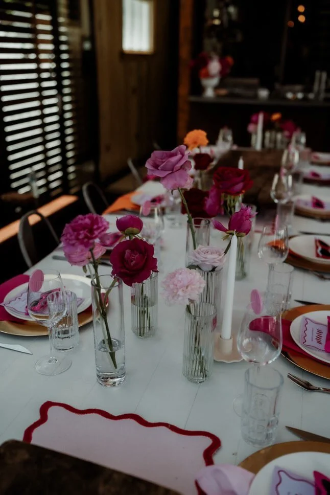 Decorated dinner table with pink and purple flowers in glass vases, candle, wine glasses, and plates set for a celebration in a cozy wood interior.