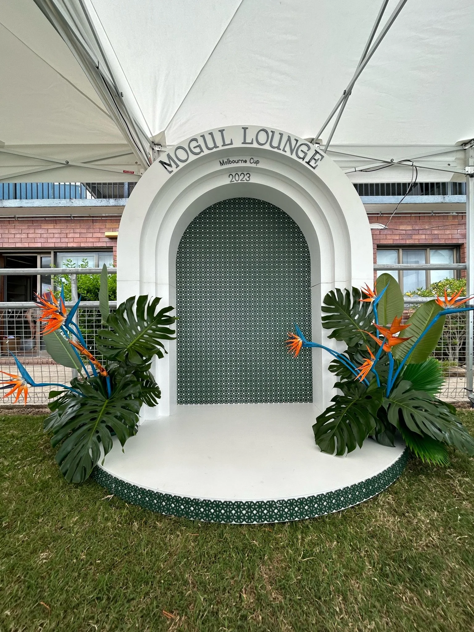 Decorative photo booth with an arch labeled 'MOGUL LOUNGE Melbourne Cup 2023,' surrounded by tropical plants with orange and blue flowers, set up on a grassy area under a white canopy.