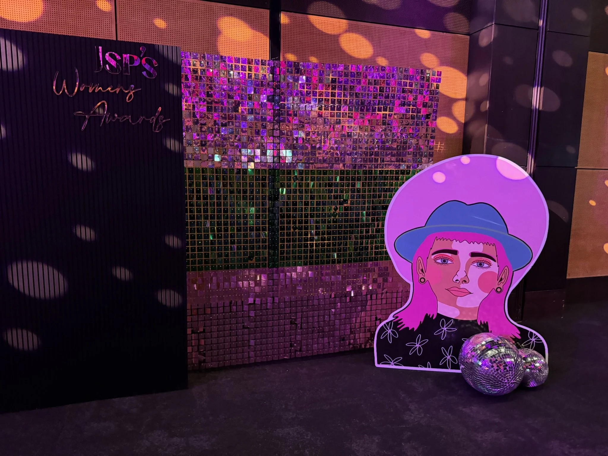 Decorative display at the ISPS Women's Awards with a sparkle backdrop, a colorful illustrated cutout of a woman with pink hair and a blue hat, and disco balls on the floor.