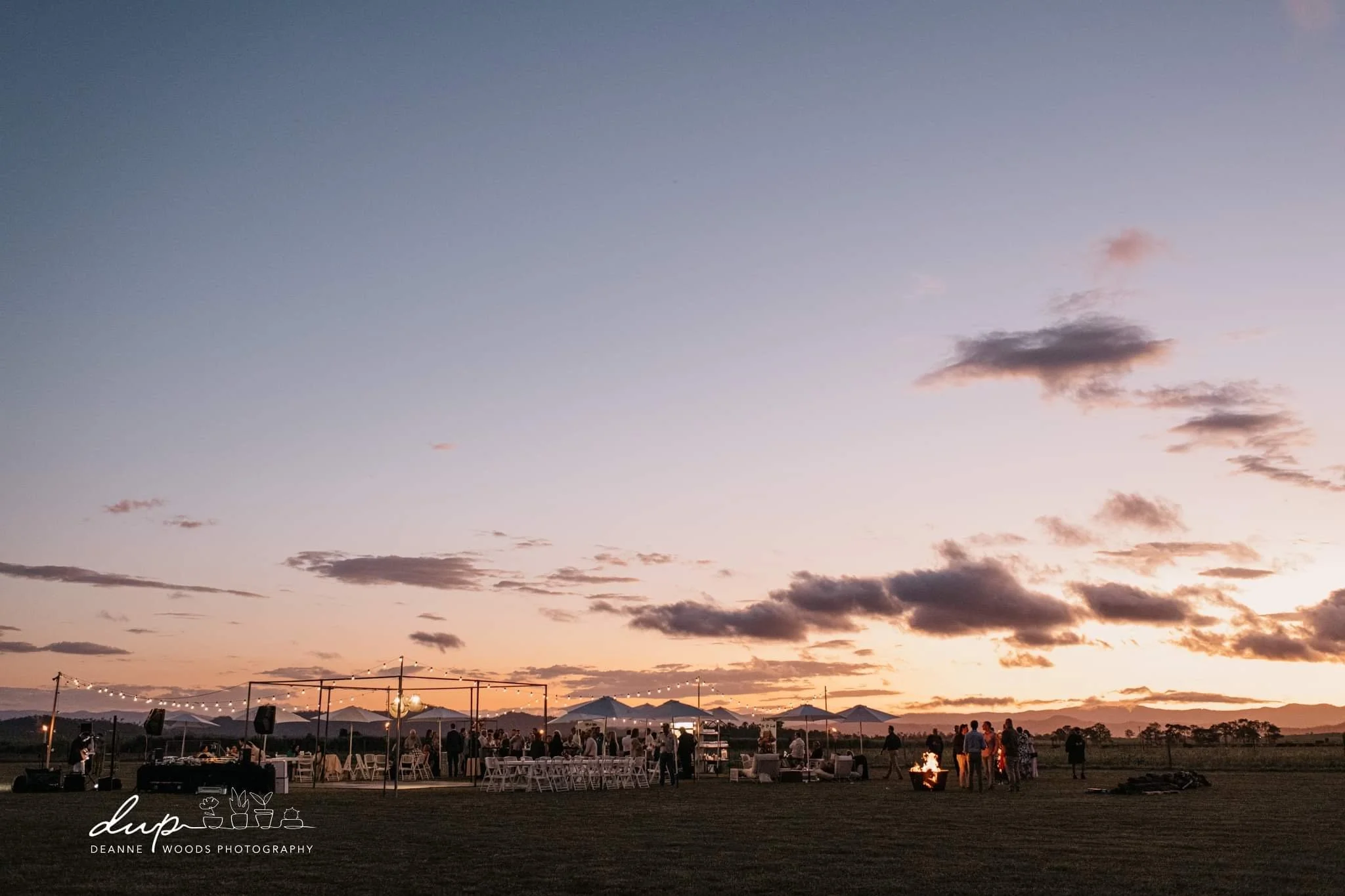 Outdoor social gathering at sunset with chairs, tables, and string lights in an open field.