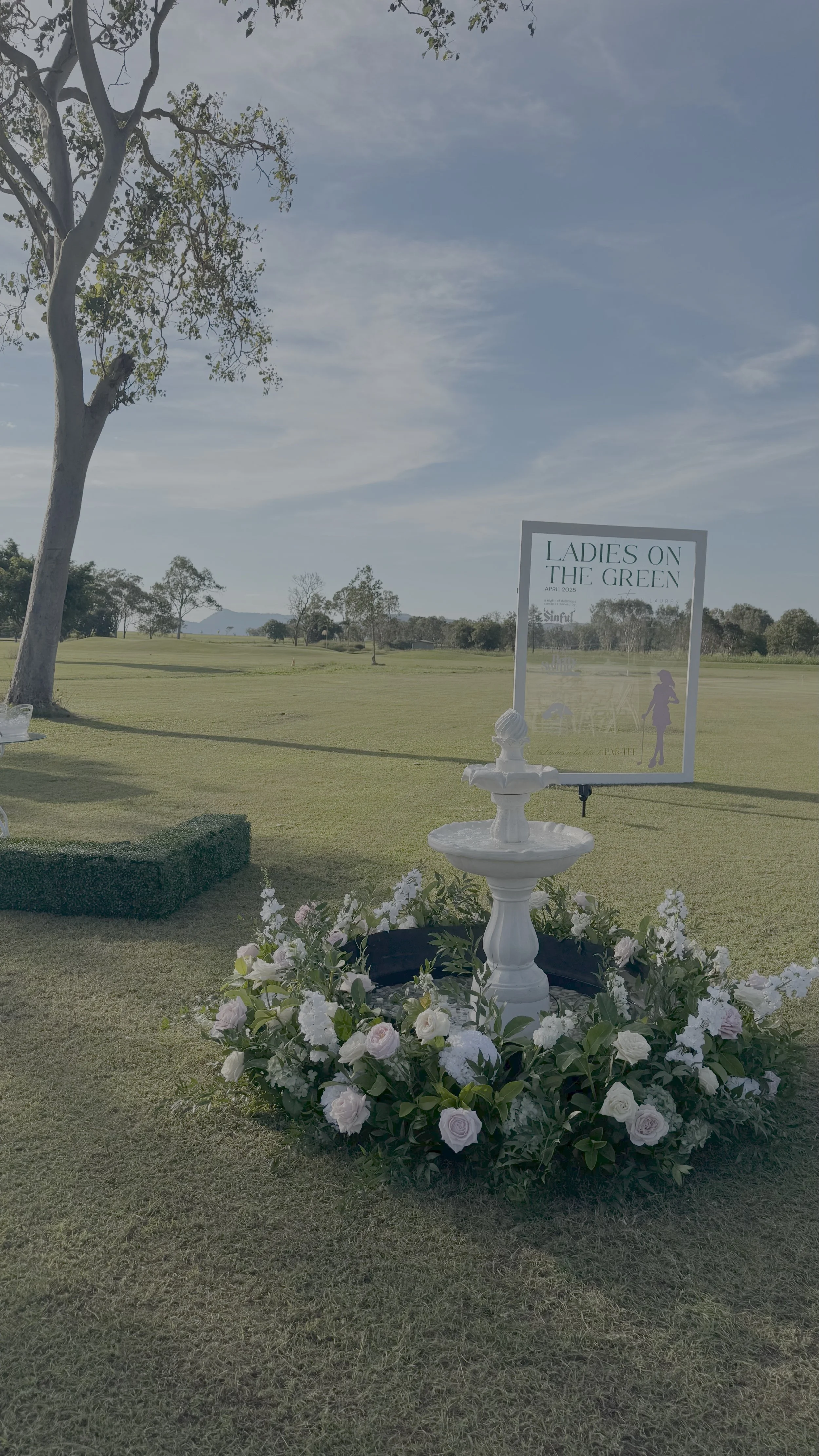 A floral arrangement surrounds a white fountain on a grassy field, with a sign reading "Ladies on the Green" and a silhouette of a woman in a hat. There are trees in the background and a blue sky overhead.