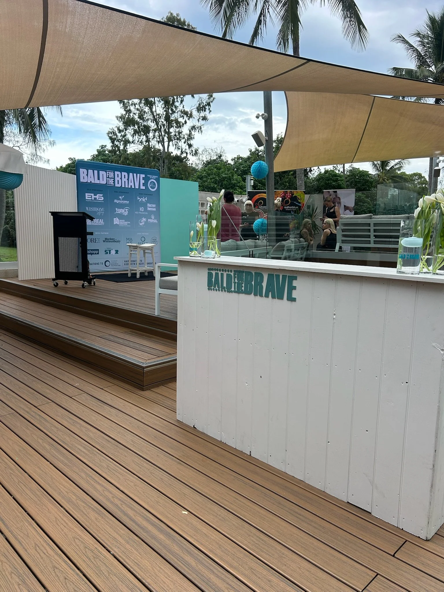 Outdoor event area with a white bar displaying the text 'Bald for the Brave,' decorated with white flowers in glass vases. There is a stage with a blue backing displaying 'Bald for the Brave' and sponsor logos, surrounded by people, some seated and s