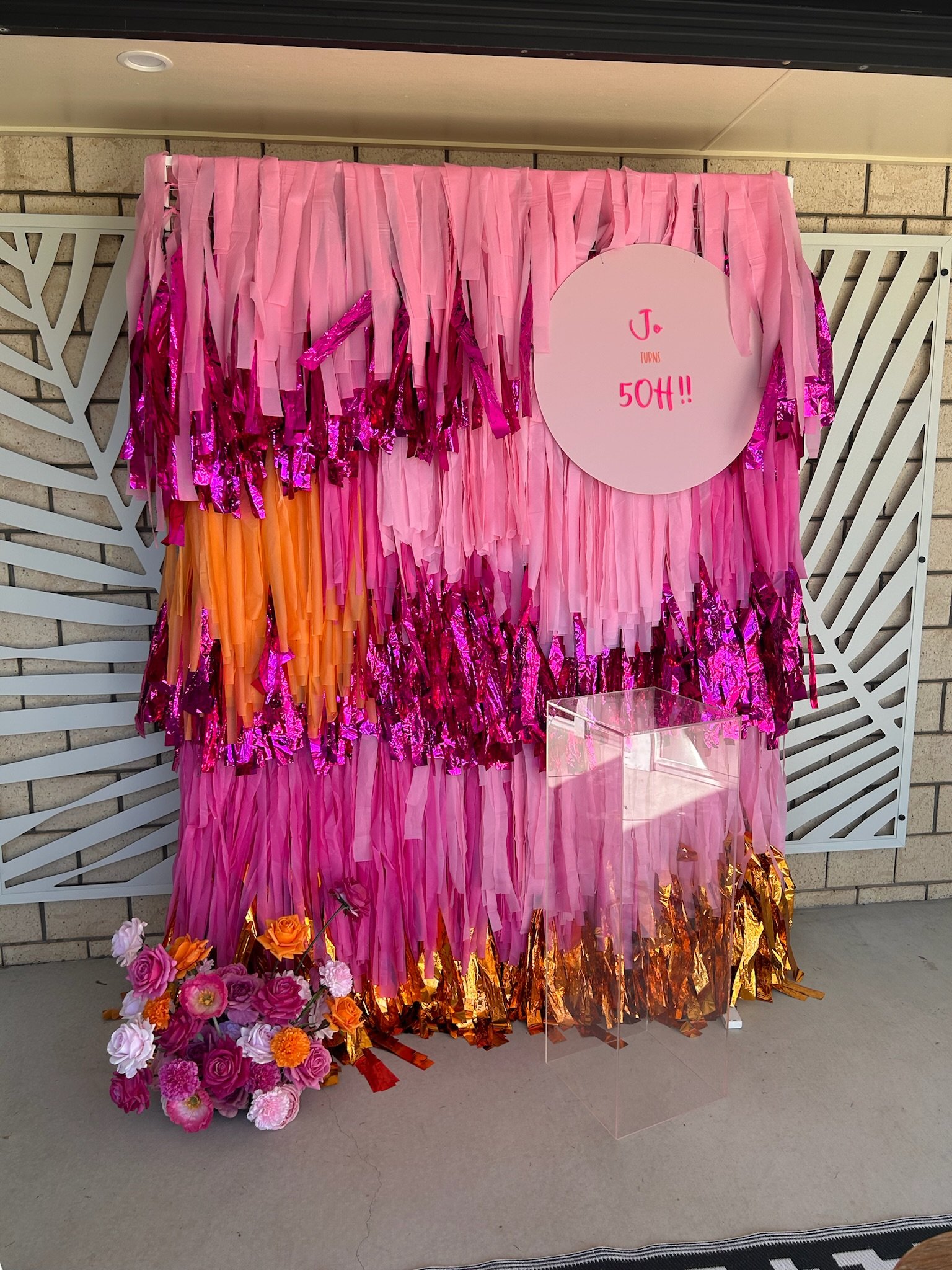 Decorative pink, orange, purple, and gold fringe backdrop with a round pink sign that says 'J. turns 50!!'. There is a bouquet of pink, purple, and orange flowers at the bottom left corner and a clear acrylic box in front of the backdrop.