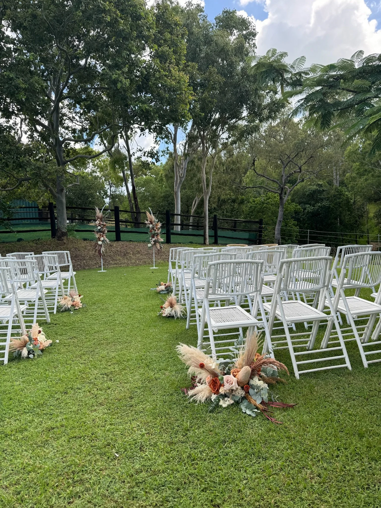 An outdoor wedding ceremony setup with white chairs arranged in rows on a grassy lawn, decorated with floral arrangements at the aisle's start and end, with a backdrop of trees and cloudy sky.