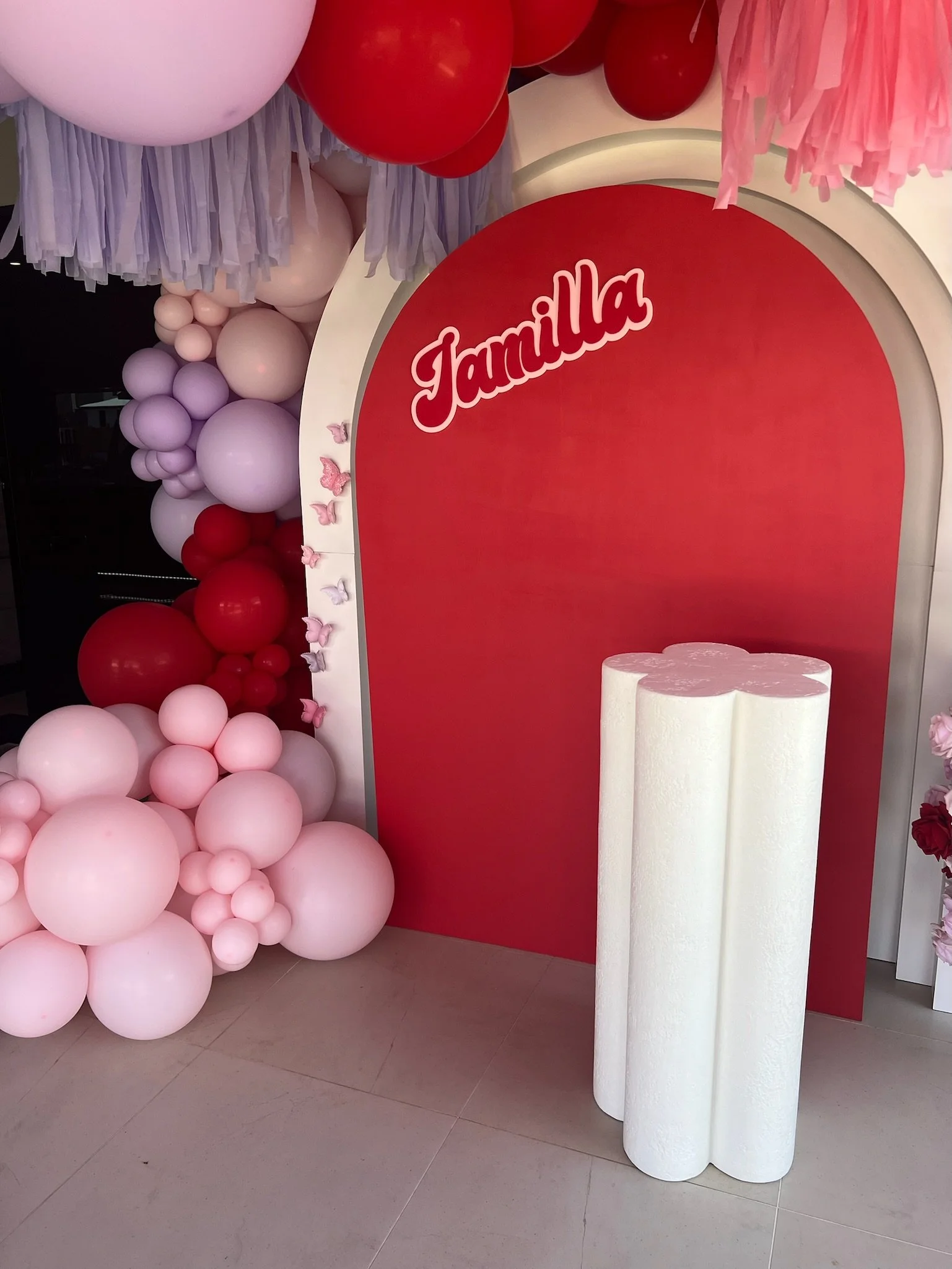 Decorative display with pink, red, and lavender balloons, a pink and purple tassel garland, and a red backdrop with the word 'Familia' in white cursive font, along with three white cylindrical pillars in front.