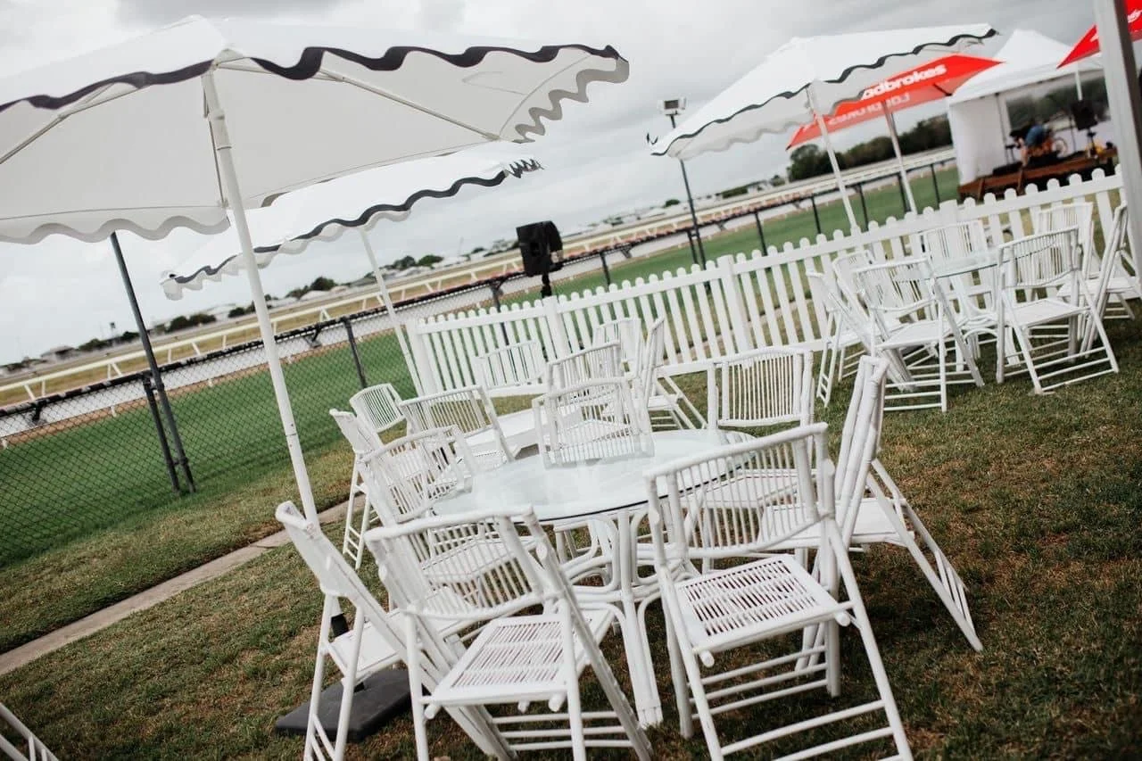 Empty outdoor seating area with white chairs and tables under large white and red umbrellas, adjacent to a racetrack or sports field with a fence and overcast sky.