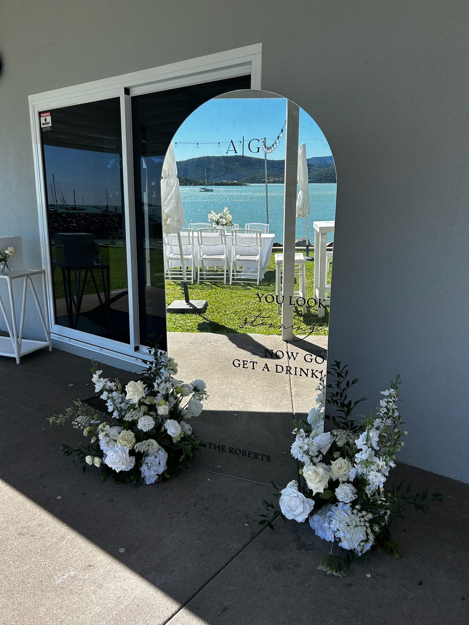 A decorative mirror with a message about a wedding, flanked by white floral arrangements, reflecting an outdoor lakeside scene with white chairs, a table, and string lights.