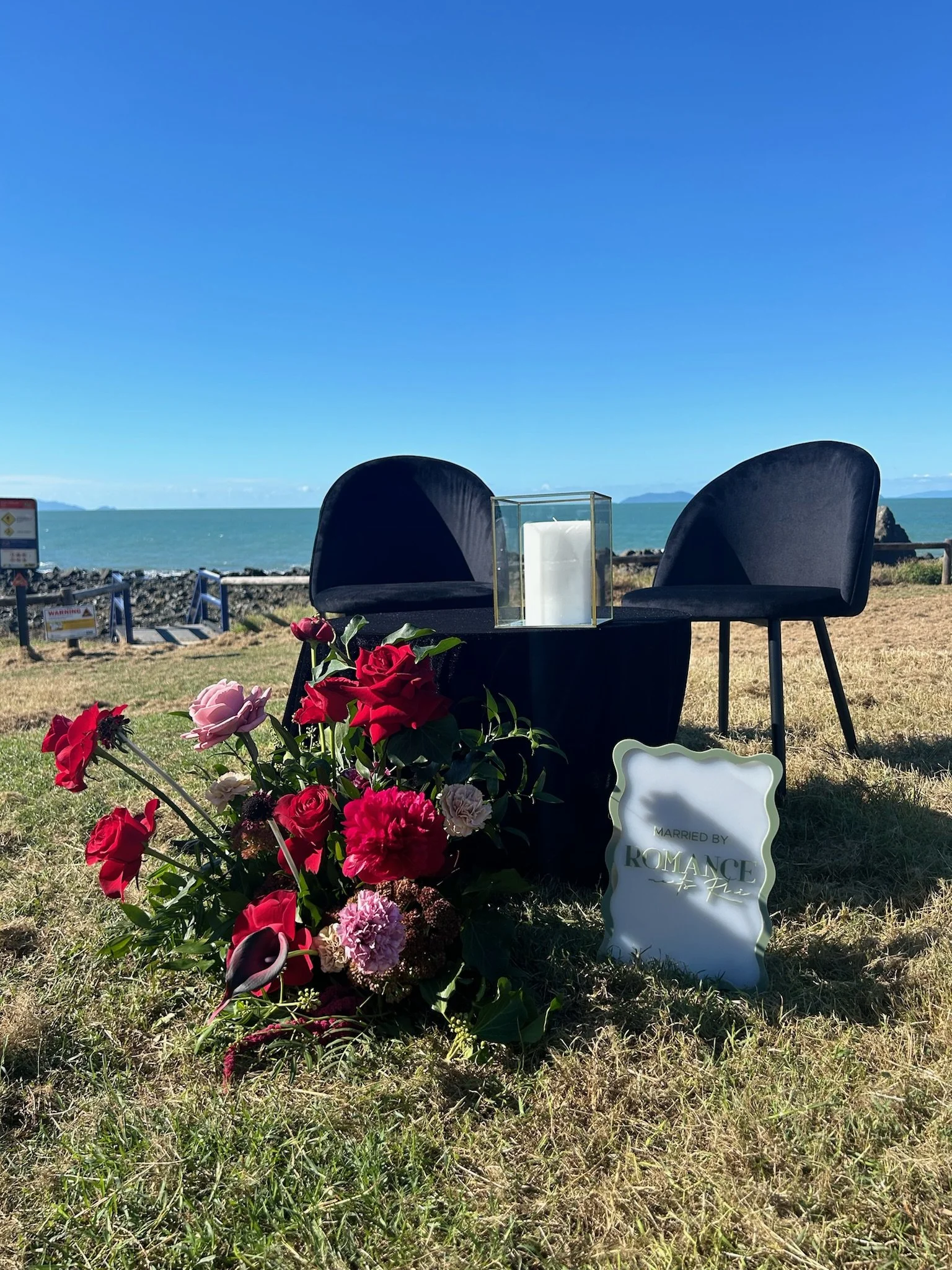 Wedding ceremony setup on a grassy area near the ocean with two black chairs, a glass candle holder, a bouquet of red and pink flowers, and a sign that reads "Married by Romance".