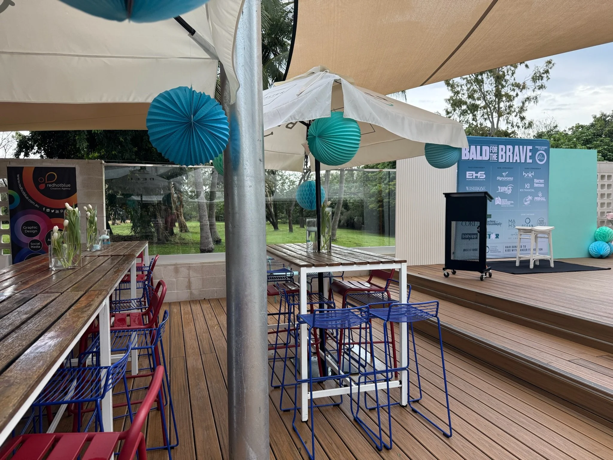Outdoor event setup with stage, chairs, tables, umbrellas, and decorative blue paper lanterns, with a sign reading 'BALD for the BRAVE'.