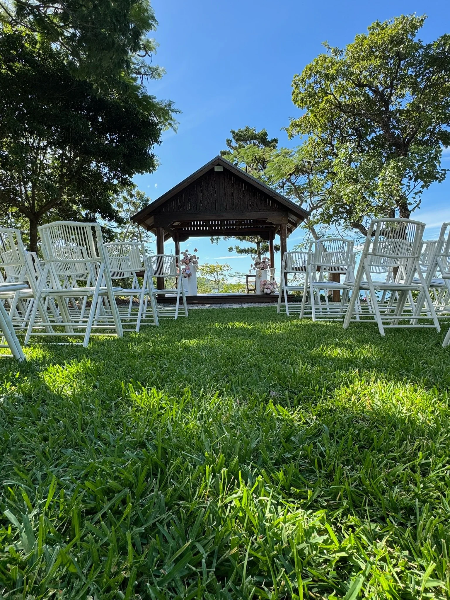 An outdoor wedding setup with white chairs arranged on a grassy lawn facing a wooden gazebo decorated with pink and white flowers, under a clear blue sky with trees on either side.