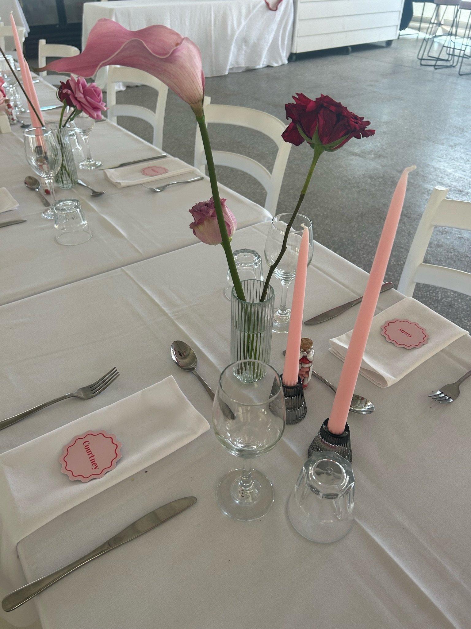 Dining table decorated with pink flowers in vases, pink candles, and place settings with napkins and cutlery for a celebration.