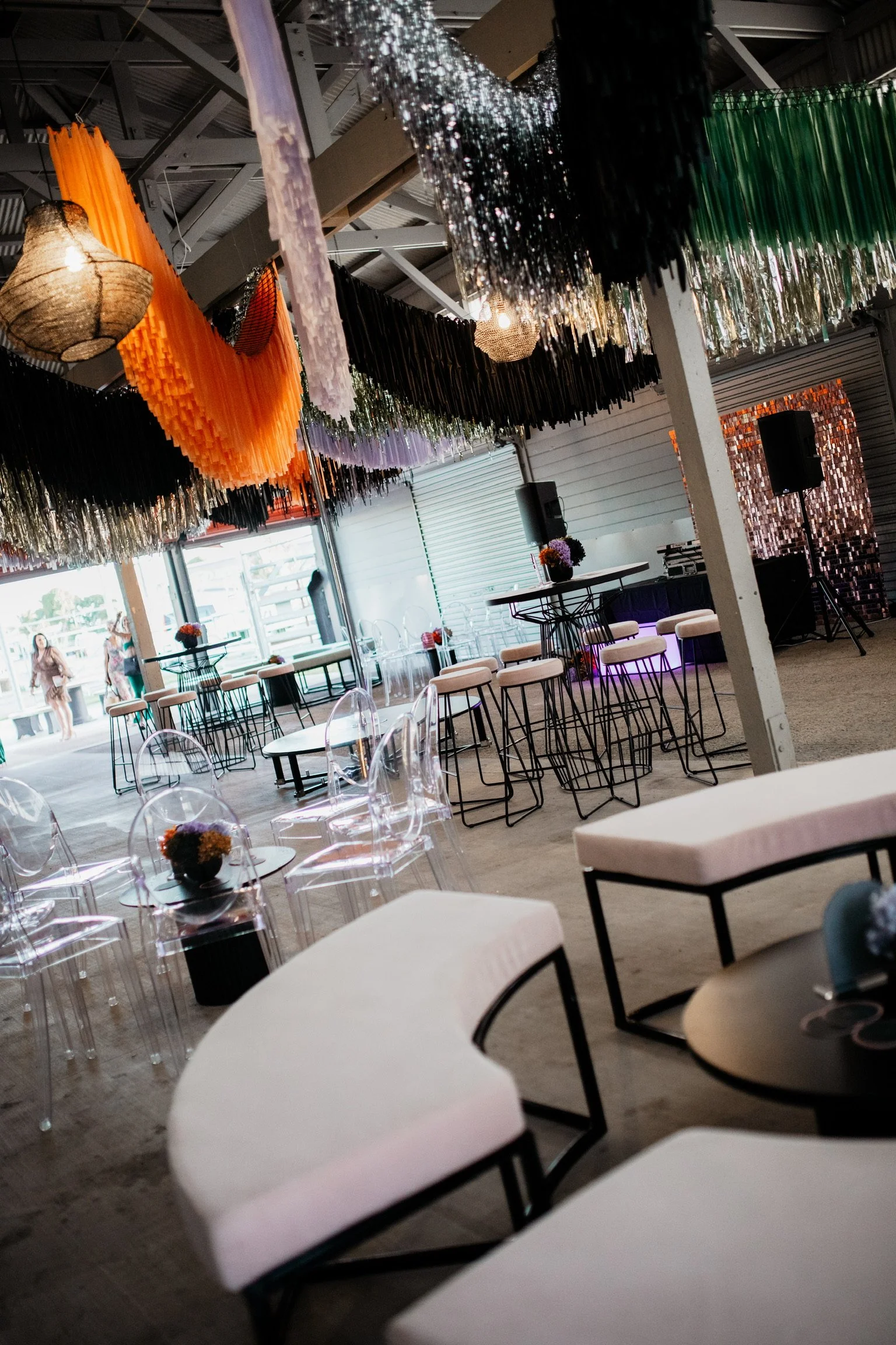 Decorated indoor event space with colorful hanging streamers, clear chairs with flower arrangements, high tables, and a DJ setup, possibly for a celebration or party.