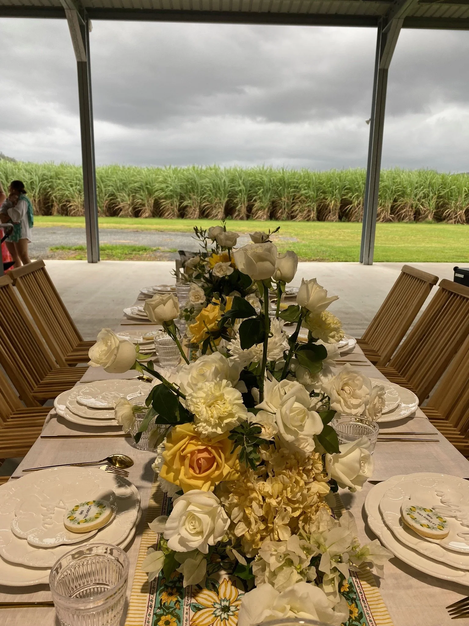 Decorated table with a floral centerpiece on a porch, overlooking sugarcane fields under cloudy sky. There are chairs, plates, glasses, and utensils set at the table.