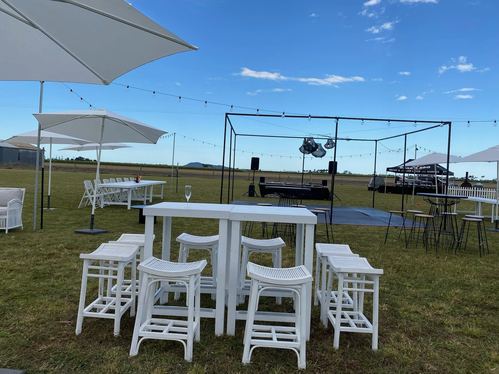 Outdoor event setup with white tables and stools, large umbrellas, a stage with lighting and musical equipment, string lights, and a clear blue sky.