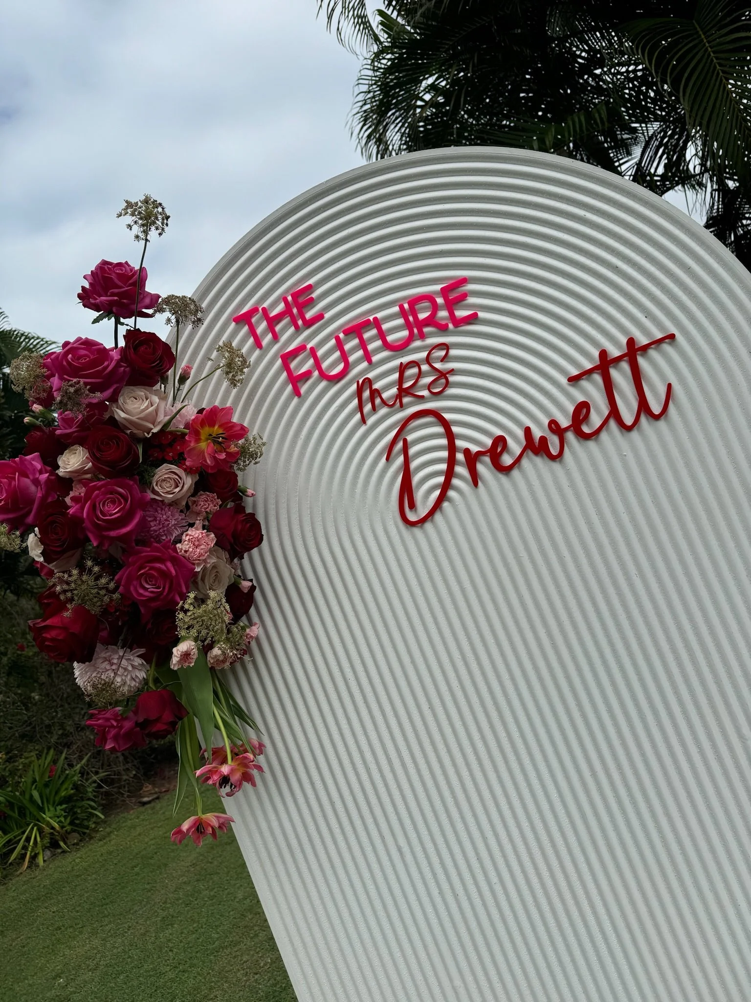 A large white sign with pink and red text that reads "The Future Mrs Drewett" and a bouquet of pink, red, and white roses with green foliage attached on the left side, outdoors with palm trees and grass in the background.