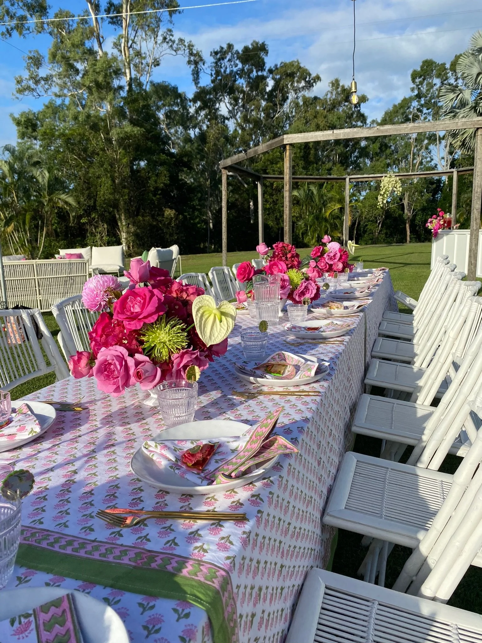 A long outdoor dining table decorated with pink and white floral centerpieces, set with plates, napkins, glasses, and gold utensils, on a grassy lawn with trees and a wooden frame structure in the background.