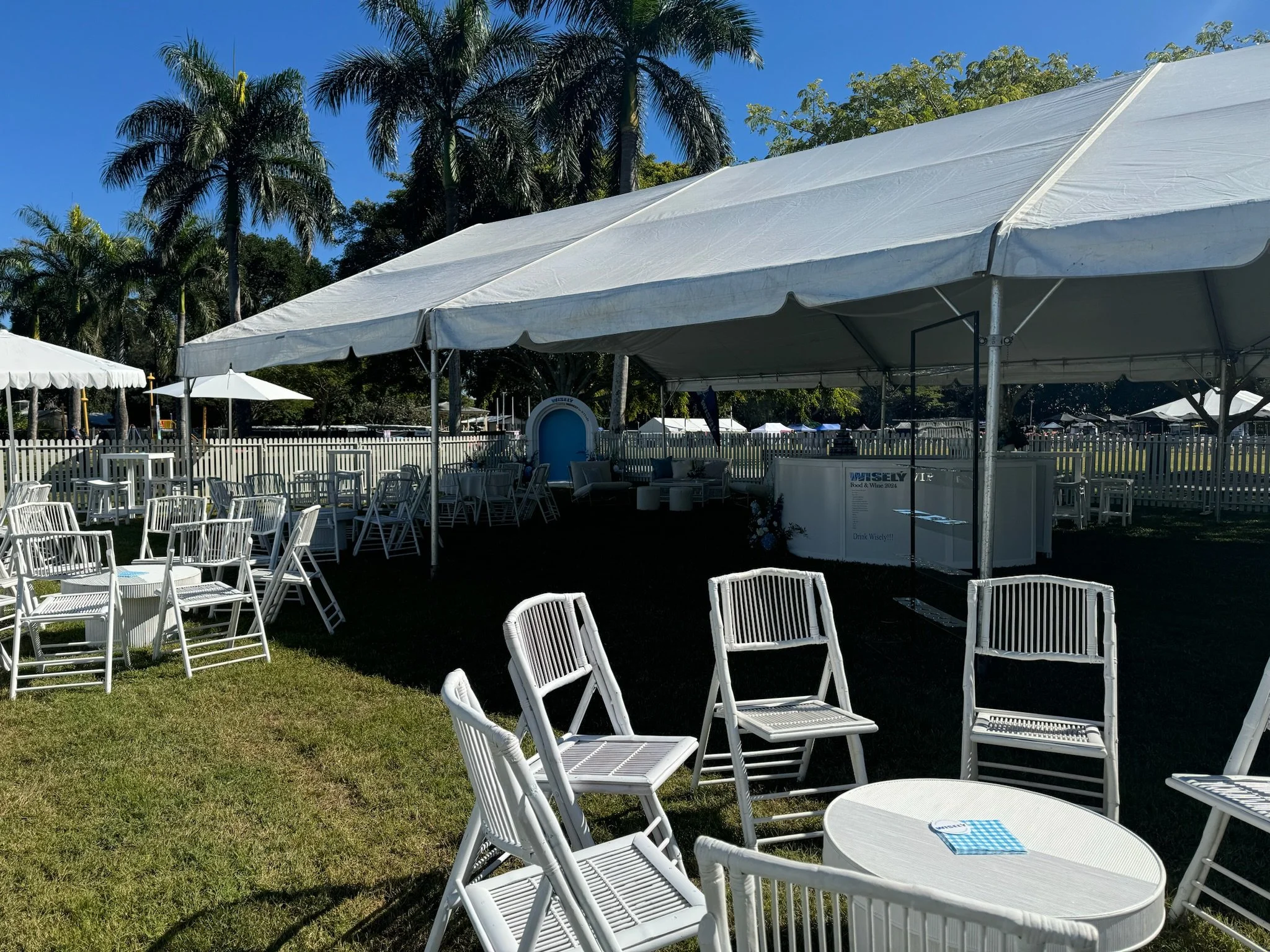 Outdoor event setup with white tables and chairs under large white tents, surrounded by palm trees and a sunny blue sky.