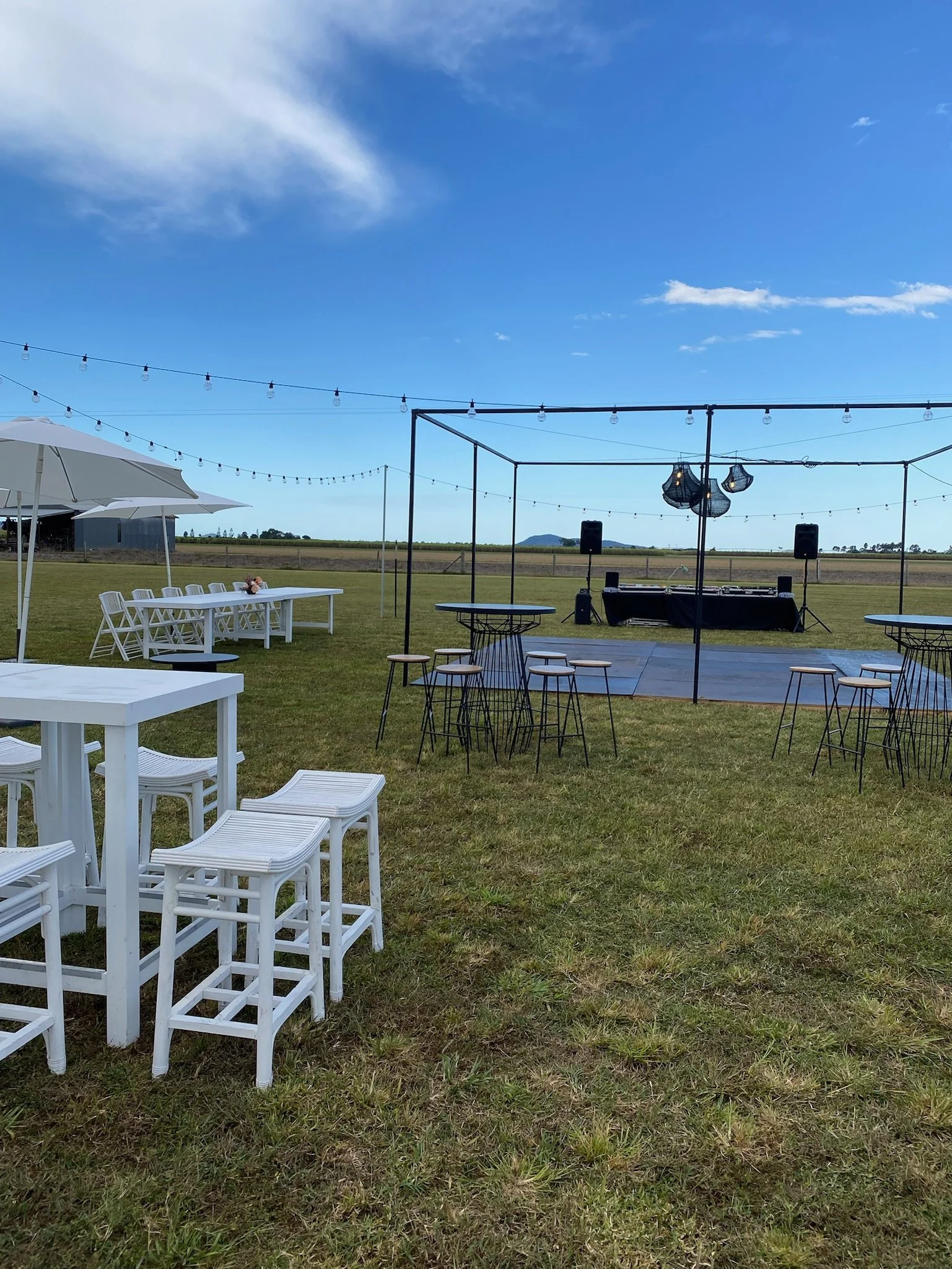 An outdoor event setup with a dance floor, speakers, and hanging lights on a clear, sunny day with blue skies and some clouds, surrounded by grass fields and distant hills.