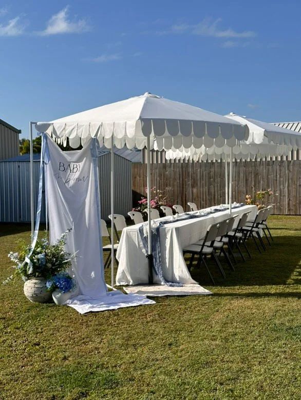 Outdoor celebration setup with long banquet table, white chairs, large white canopy, floral decorations, and a banner that says 'Baby'. 