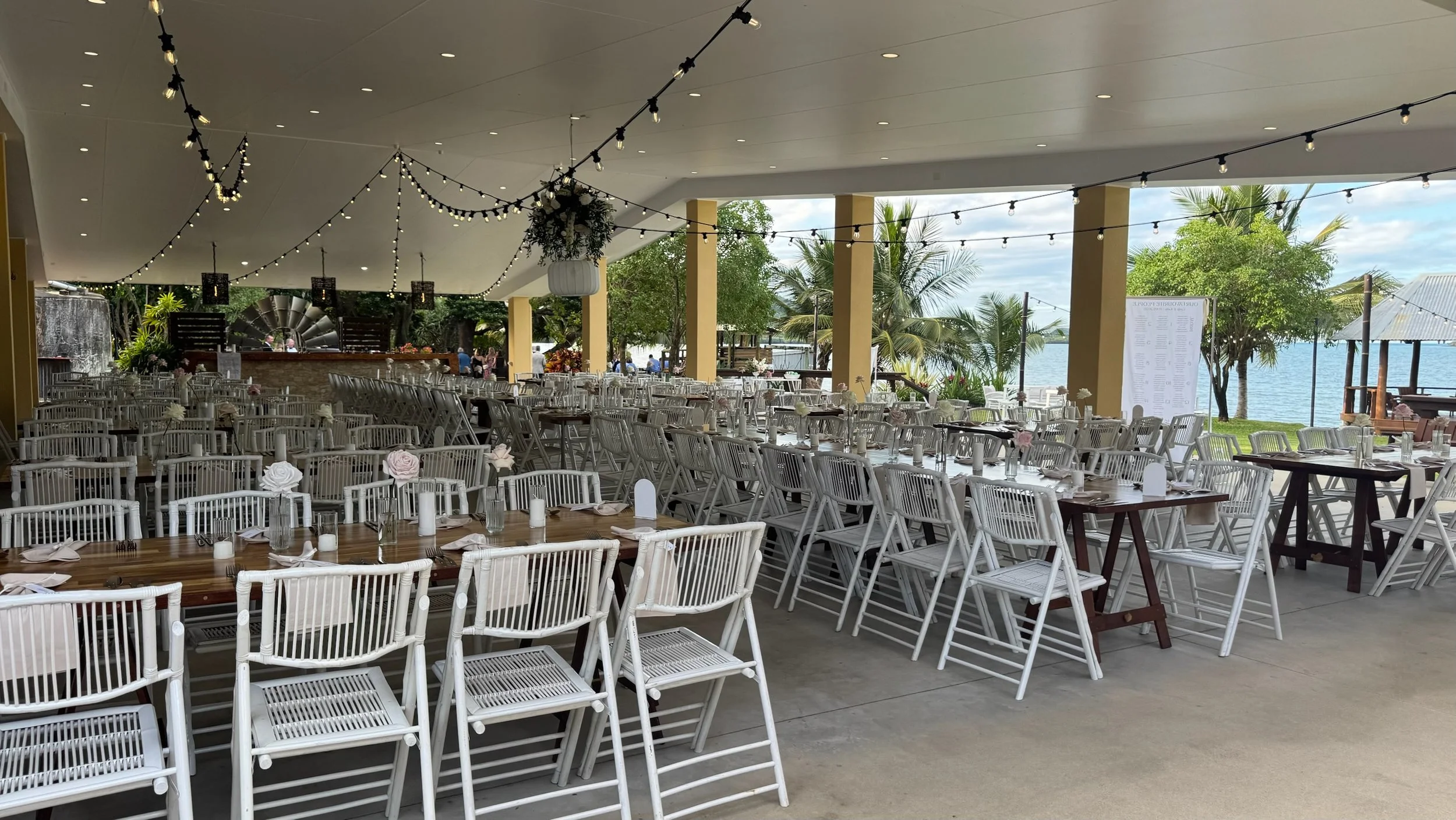 Outdoor wedding or event venue with white chairs and tables arranged under a covered patio near water, decorated with string lights and flowers.
