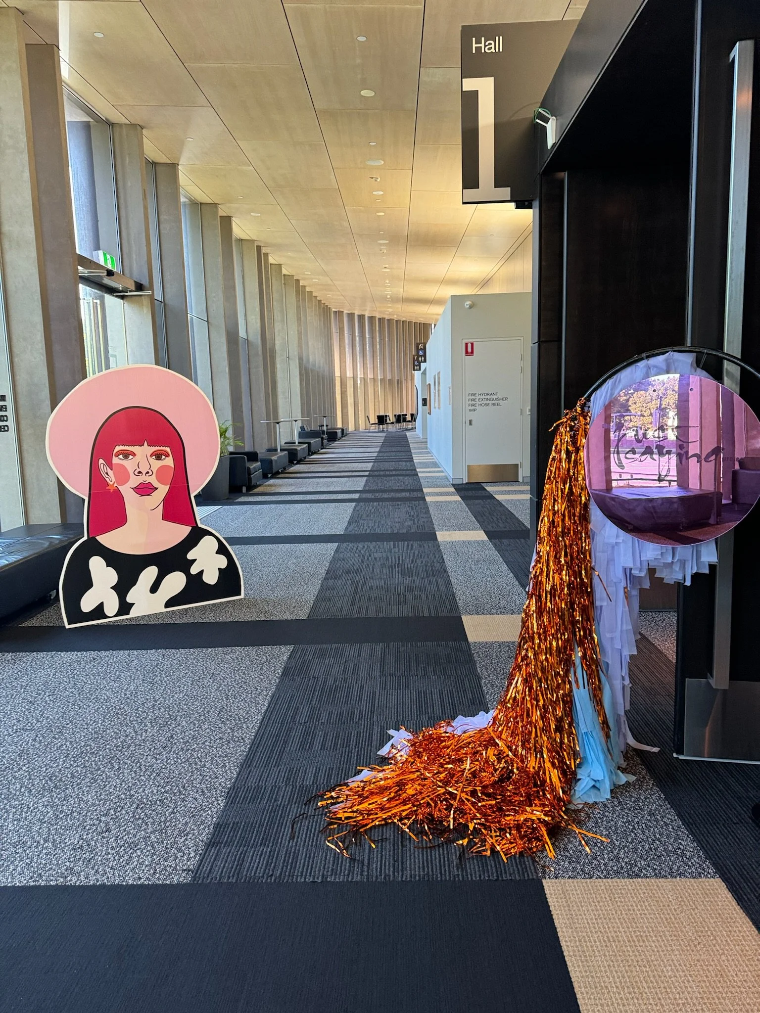A modern, spacious hallway with a high ceiling and large windows on the left. There is a cardboard cutout of a woman with red hair and a pink hat on the left side. On the right side, there is a decorative piece made of orange tinsel and white fabric.