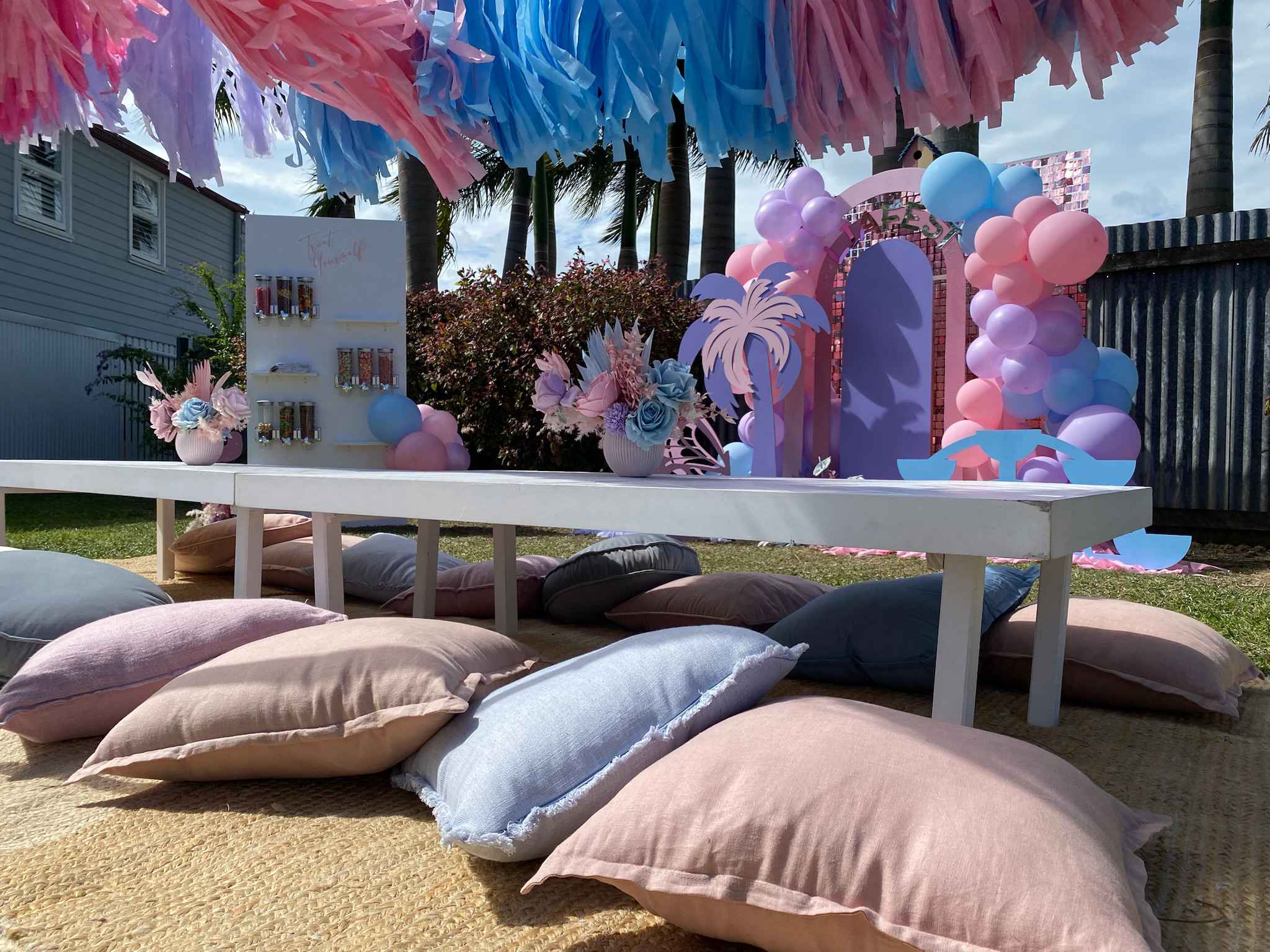 Decorative outdoor party setup with pastel pink, purple, and blue balloons, cushions on the ground, a white table with flower vases, and a pink arch with the word 'HAPPY' surrounded by palm tree and tropical-themed decorations.
